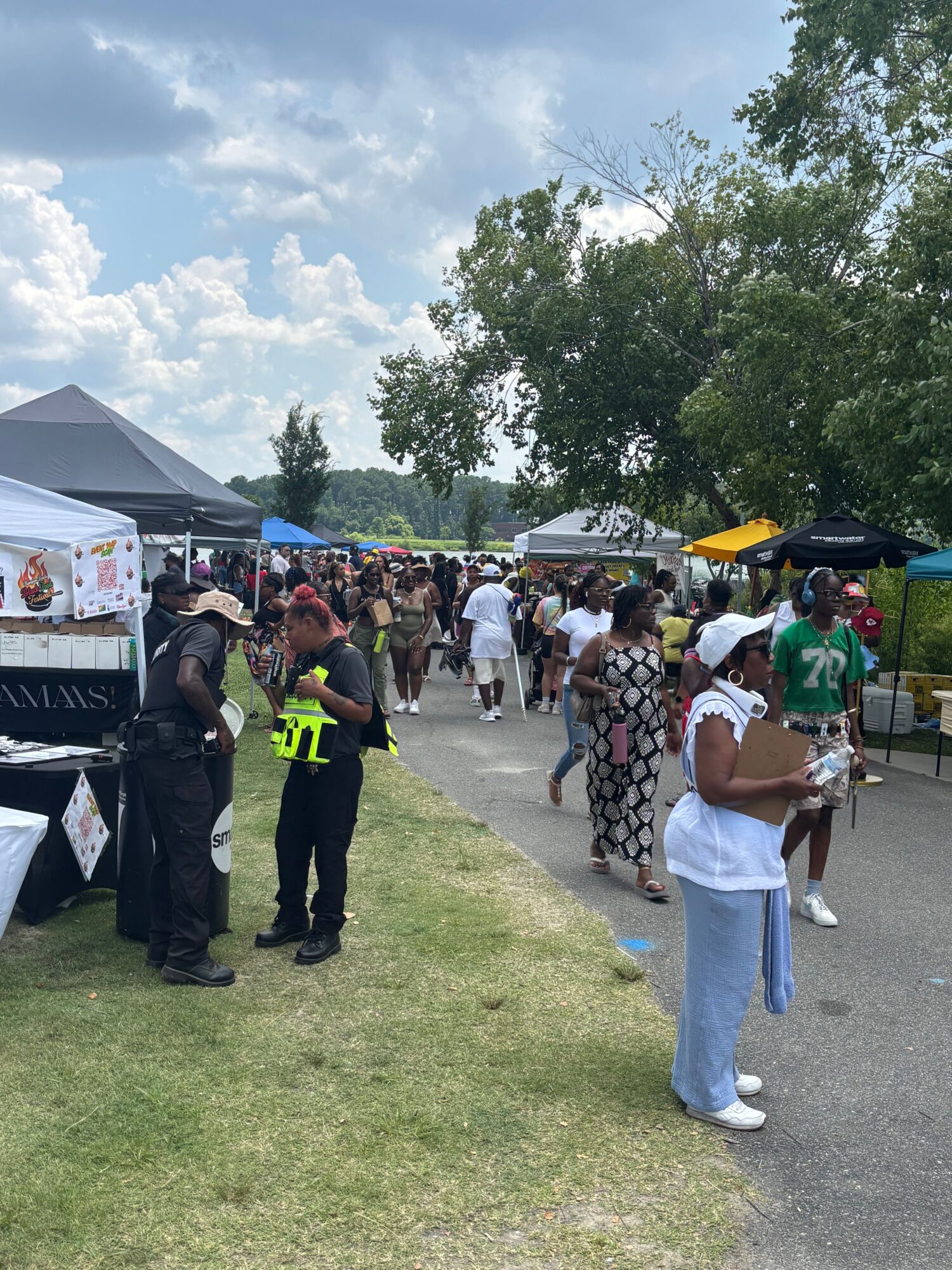 People gather outdoors near tents and trees, some wearing masks, with a police officer present, on a sunny day.