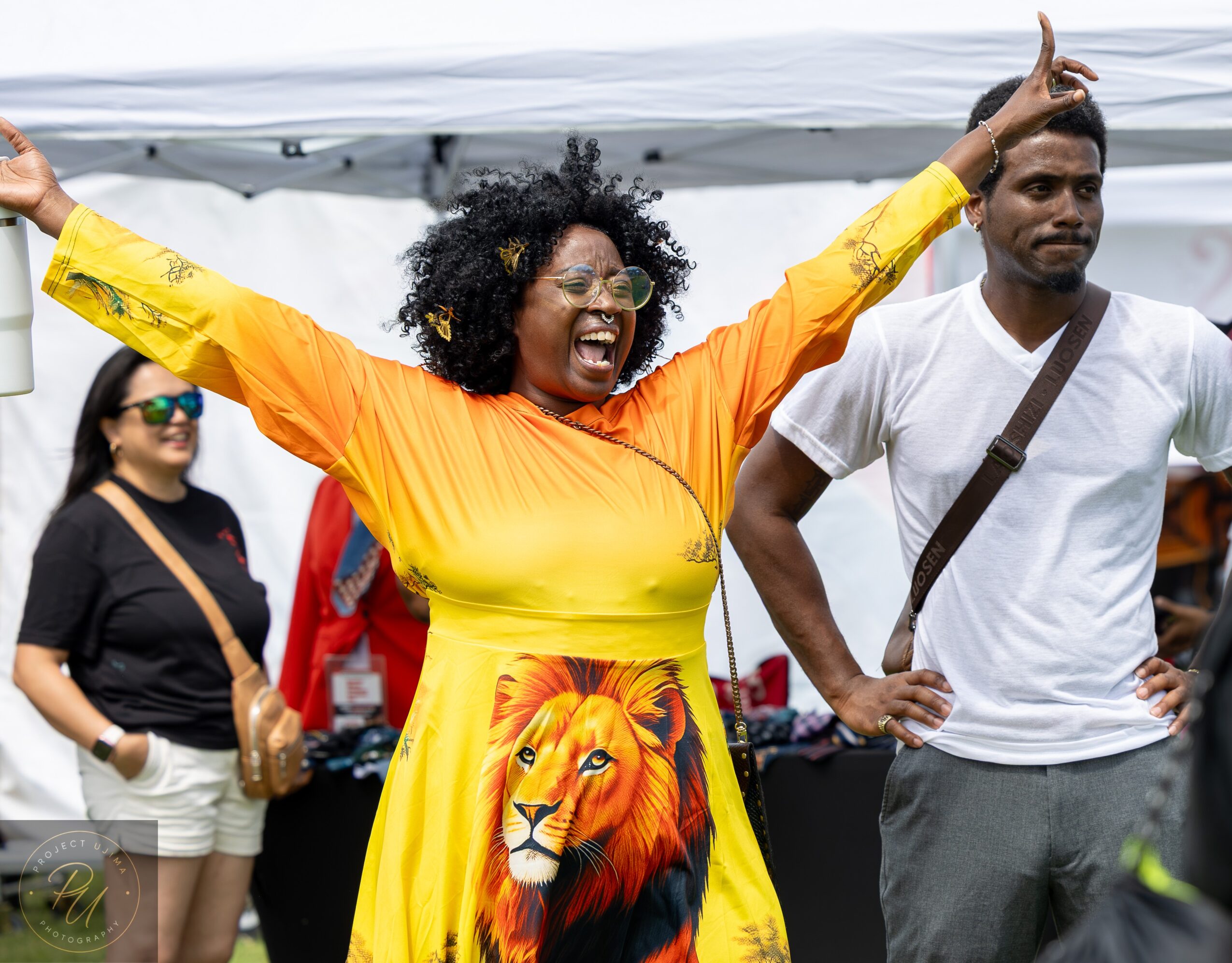 Woman with curly hair wearing a yellow dress with a lion print, smiling and raising her arms, outdoors with people around.