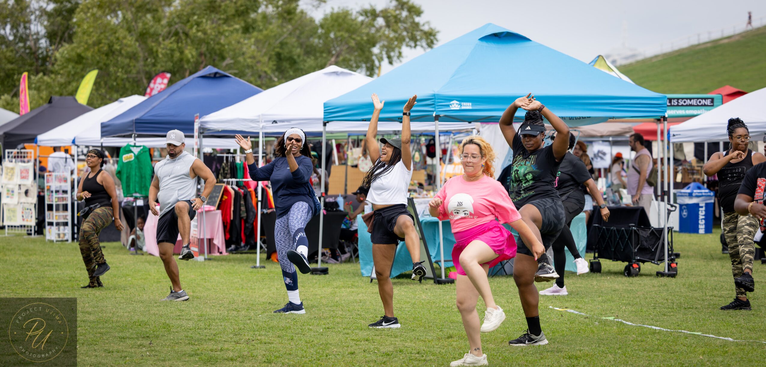 Group of people dancing outdoors on grass with tents and trees in background, some wearing casual clothing and hats.