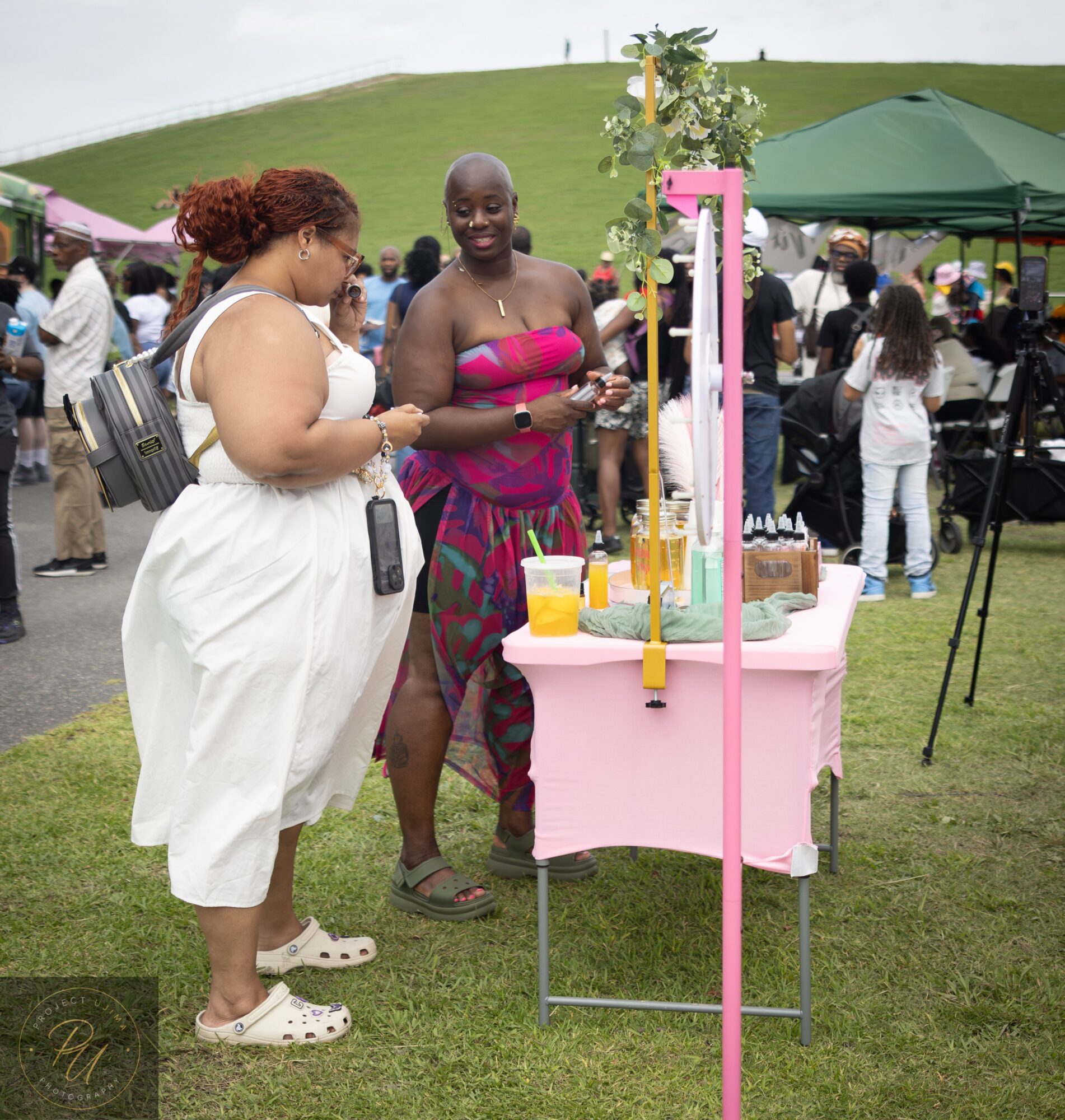 Two women at an outdoor event, one in a white dress and the other in a colorful strapless dress, near a pink display table.