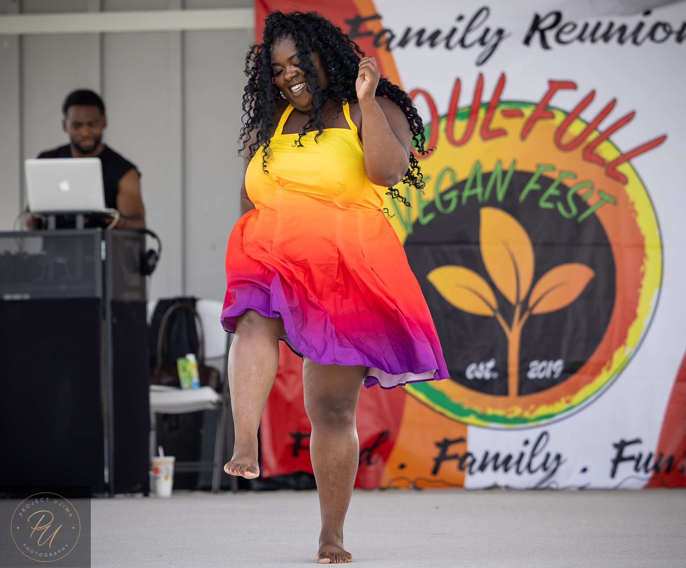 Woman dancing in colorful dress on stage with a man at a DJ booth in background, festival banner behind her.