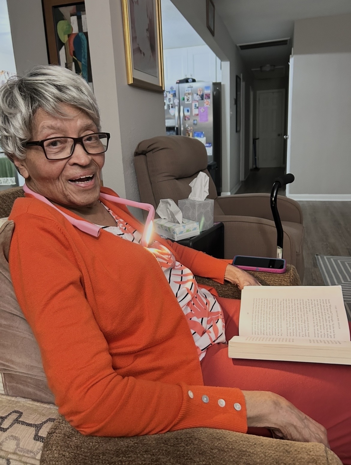 Older woman with glasses and short gray hair sitting on a couch, smiling, with an open book on her lap.