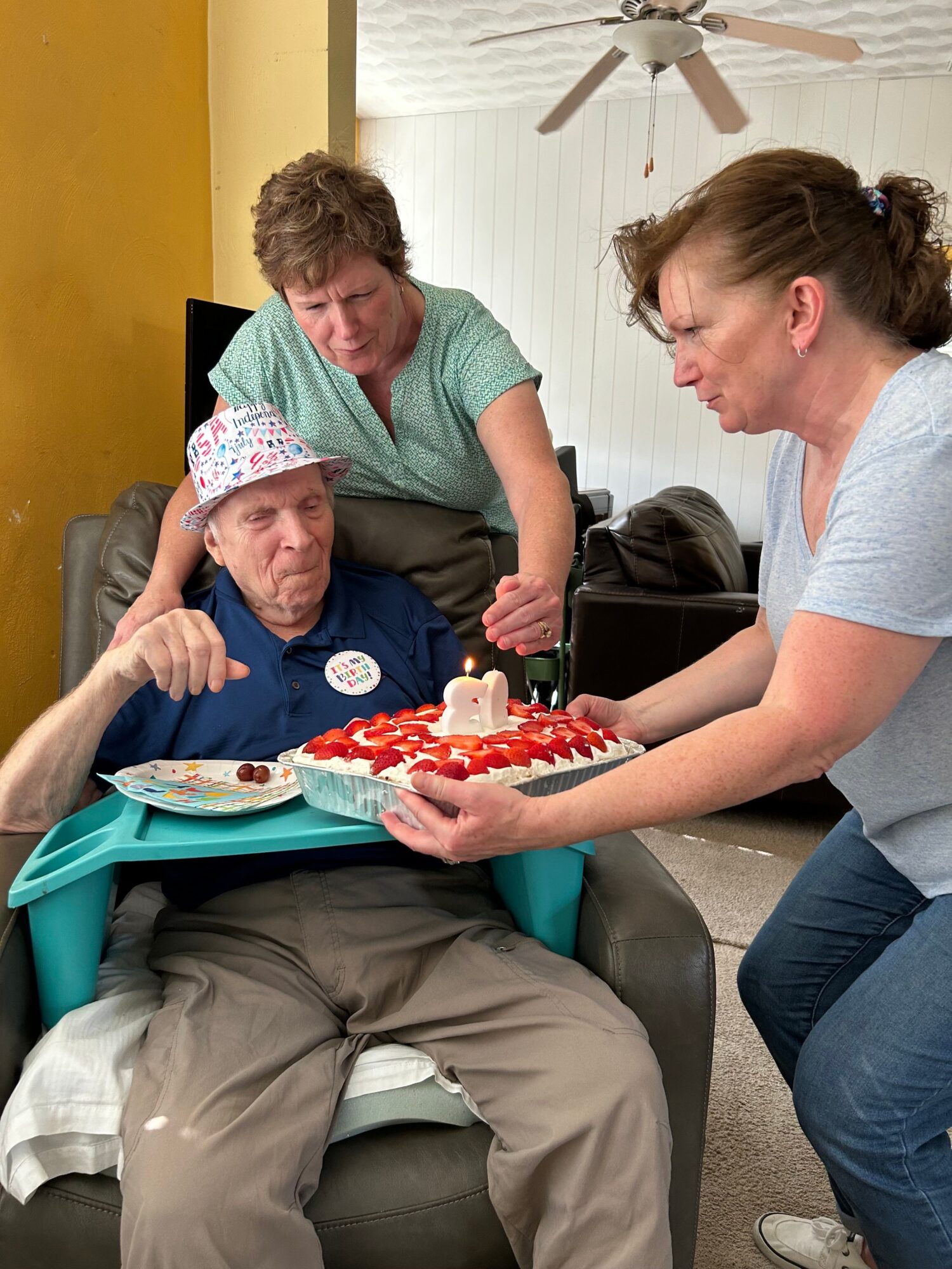 Two women and a child sitting on a couch, with one woman holding a plate of cake and the child giving a thumbs-up.