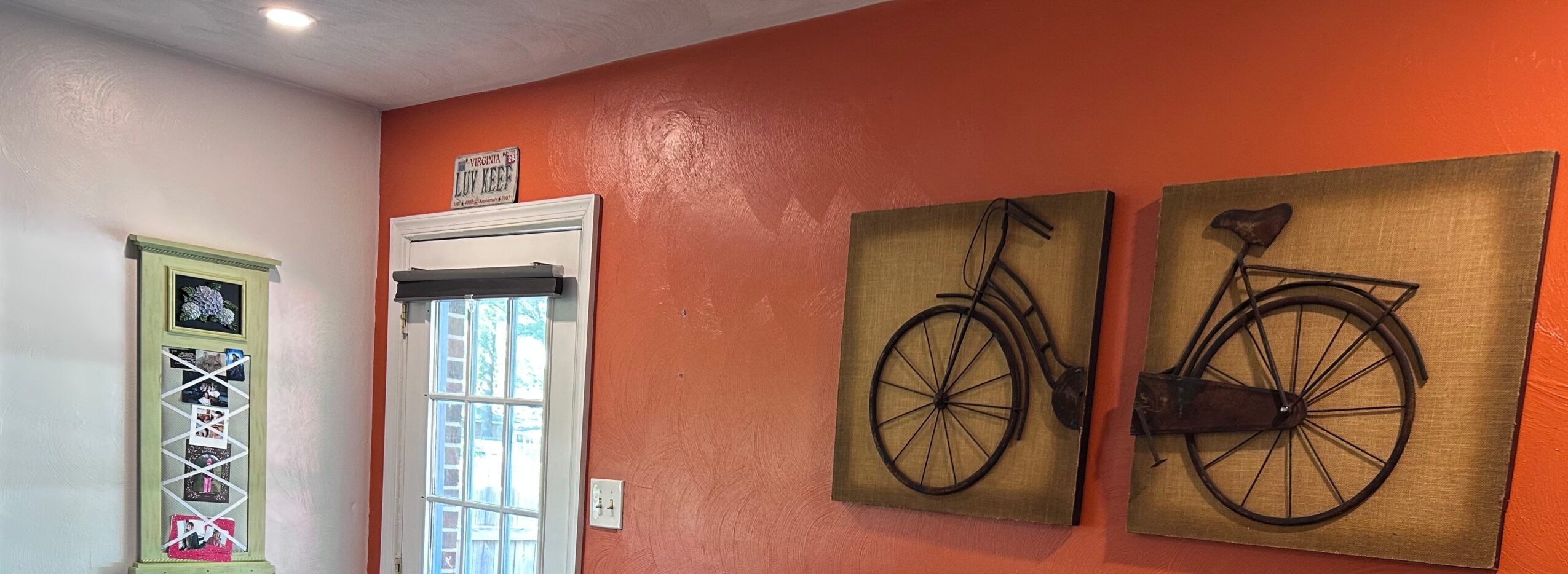 Two wall art pieces of bicycles hang on a red wall in a room with a window and a green cabinet.