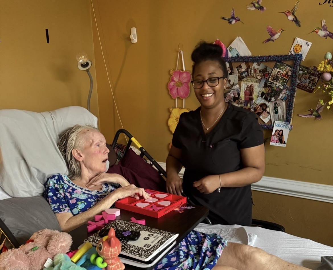 A woman in bed with a floral shirt interacts with a smiling caregiver in a black uniform in a room with yellow walls and colorful decorations.