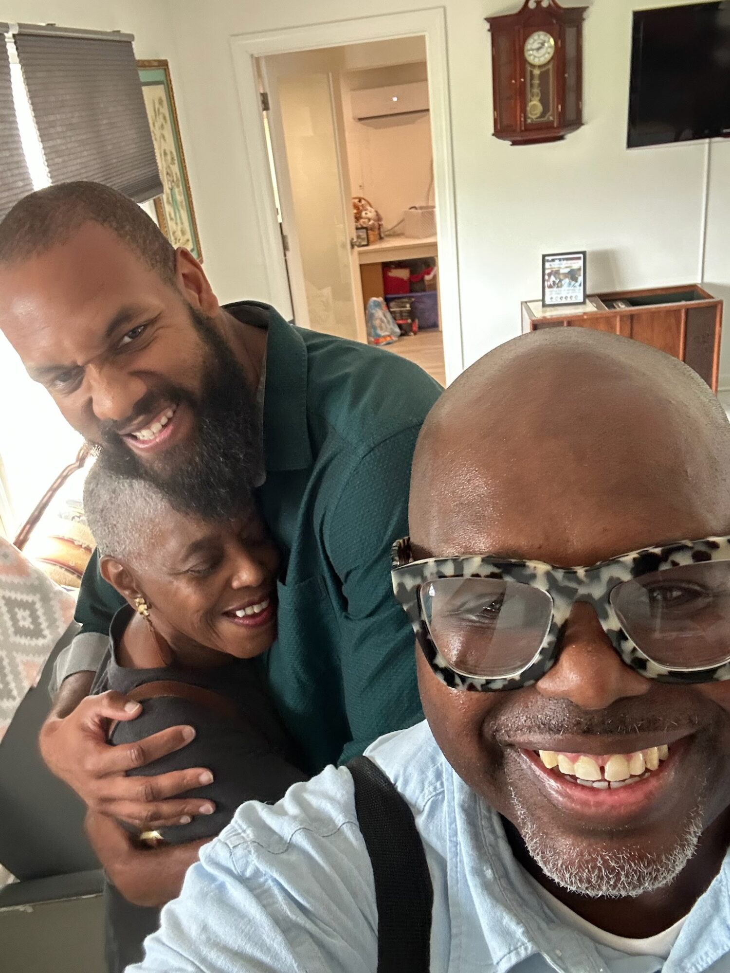 Three people smiling and hugging indoors, with a man wearing glasses in the foreground, a woman and man behind him.