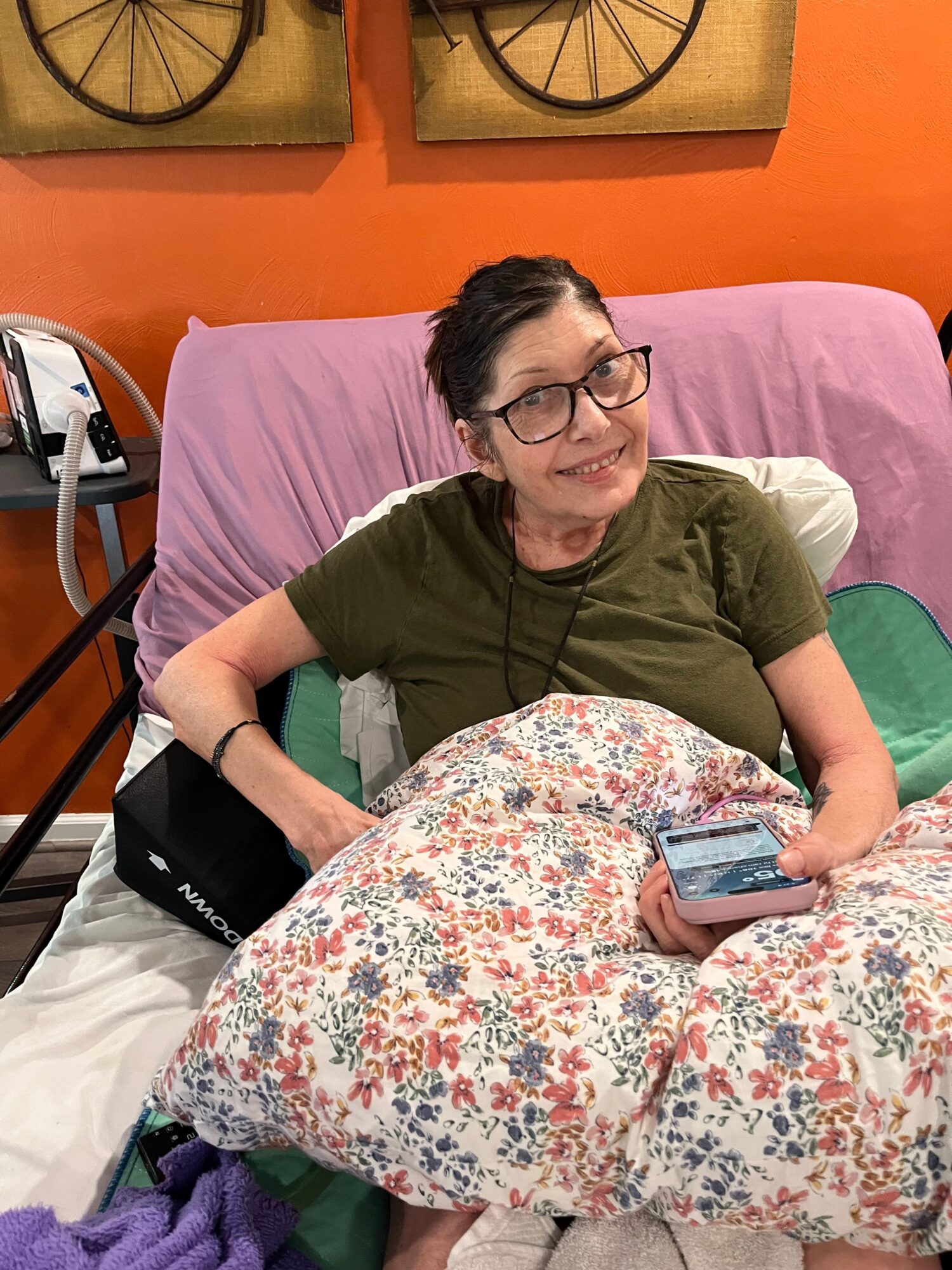 Person with glasses and dark hair, wearing a green shirt, sitting on a bed with floral bedding, holding a phone.
