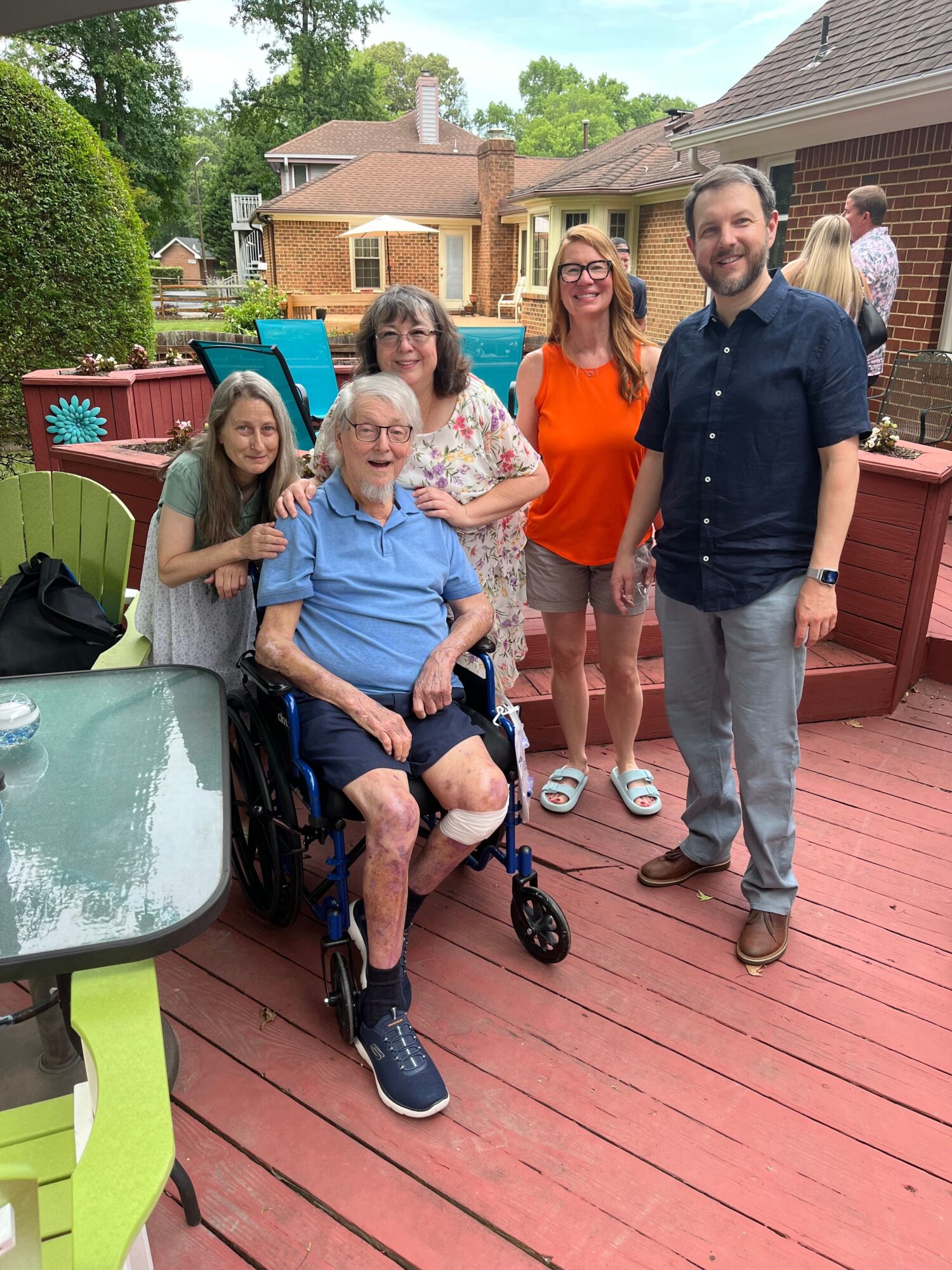 Group of six people outdoors on a wooden deck, some sitting and some standing, with trees and houses in background.