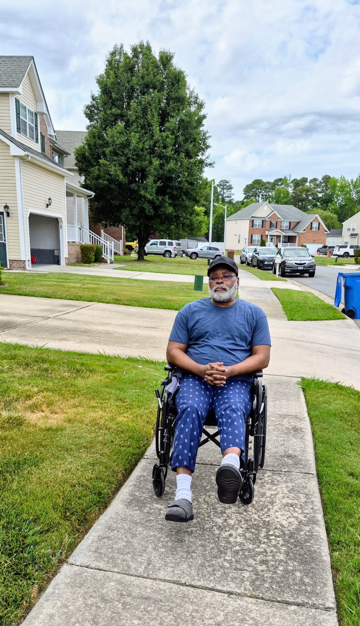 Person in a wheelchair sitting on a sidewalk in a residential neighborhood with houses, trees, and parked cars in the background.