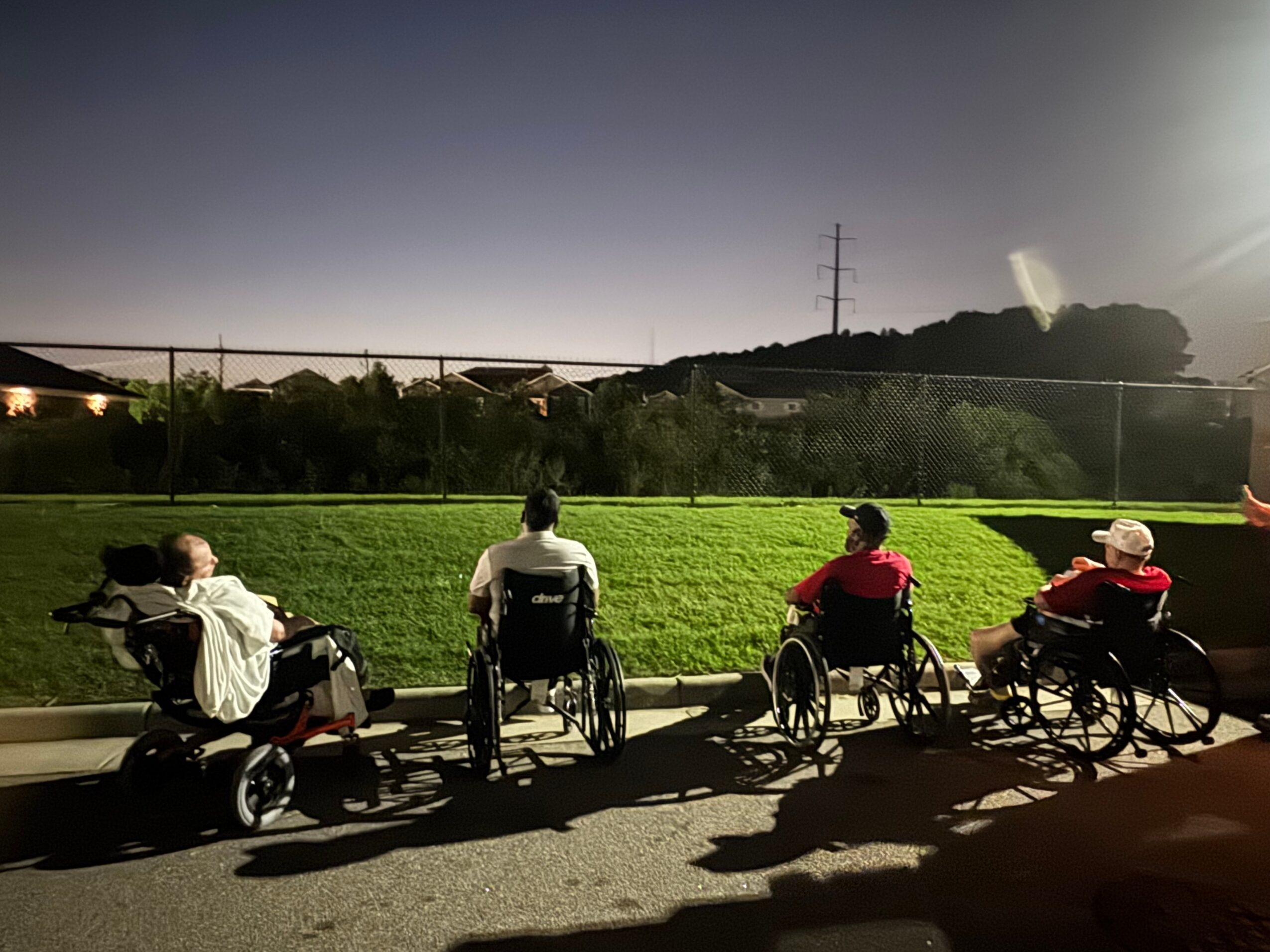 Four people in wheelchairs sit on a sidewalk facing a grassy field with trees and power lines in the background.