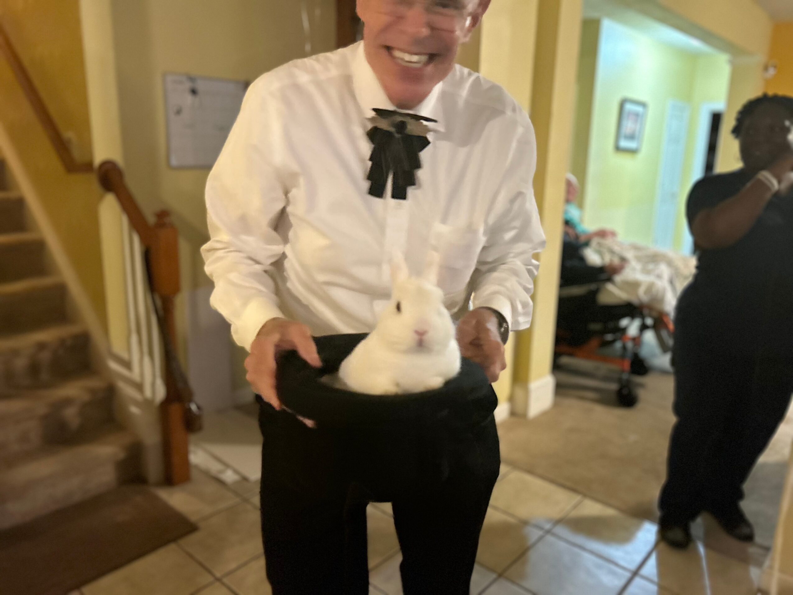 Person in white shirt holding a white rabbit indoors, smiling, with a staircase and another person in the background.