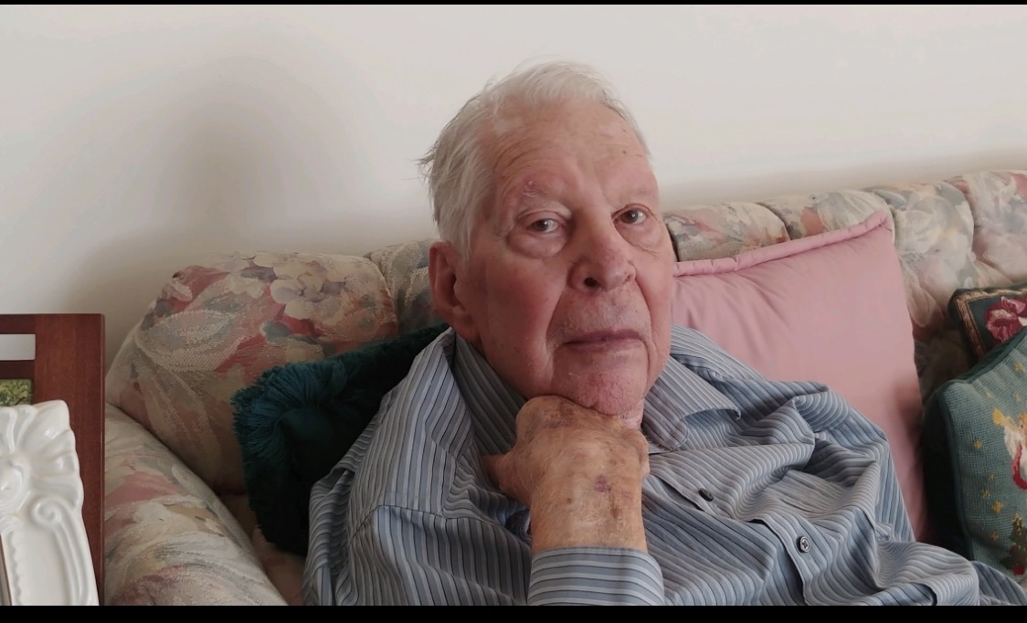 Elderly man with white hair sitting on a sofa, resting his chin on his hand, wearing a striped shirt.