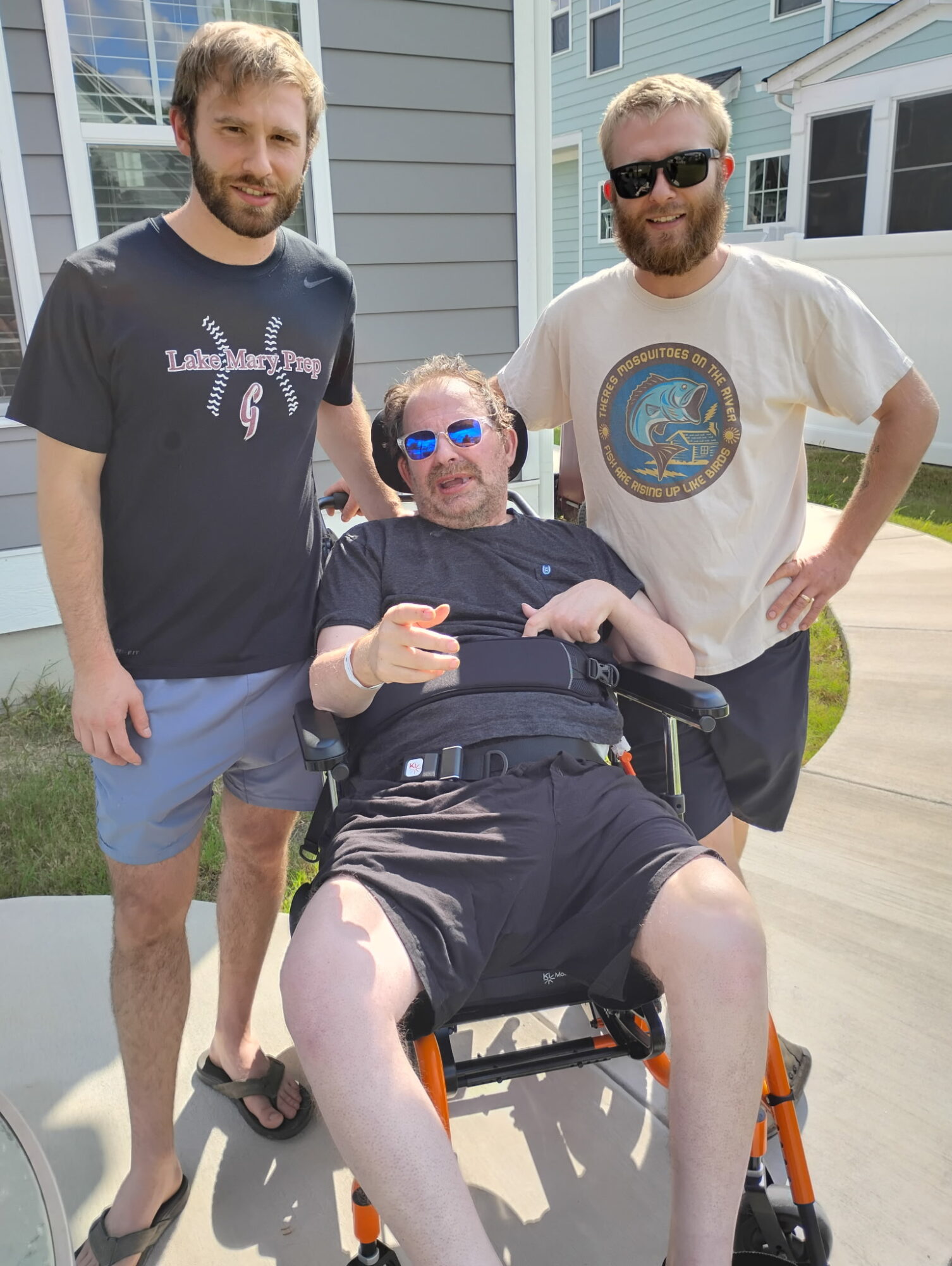 Three men outdoors, one in a wheelchair, two standing, all smiling, in front of houses, sunny day.