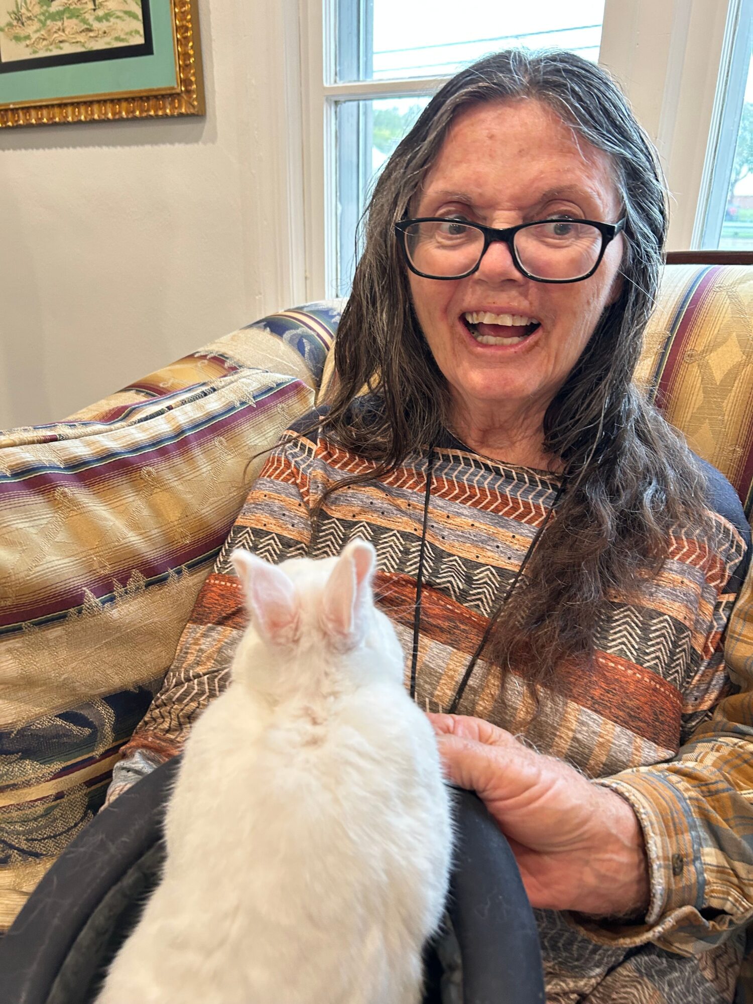 Woman with glasses smiling at a white cat indoors, window in background, colorful striped shirt, holding the cat gently.