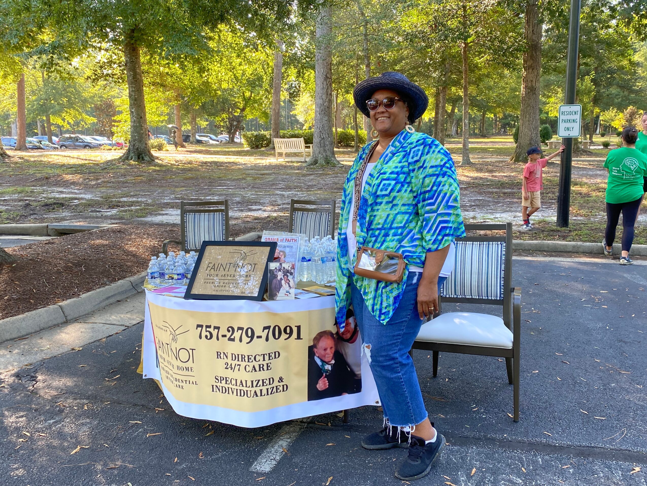 Woman in blue patterned shirt and hat standing next to a table with a sign, in a park with trees and people in background.