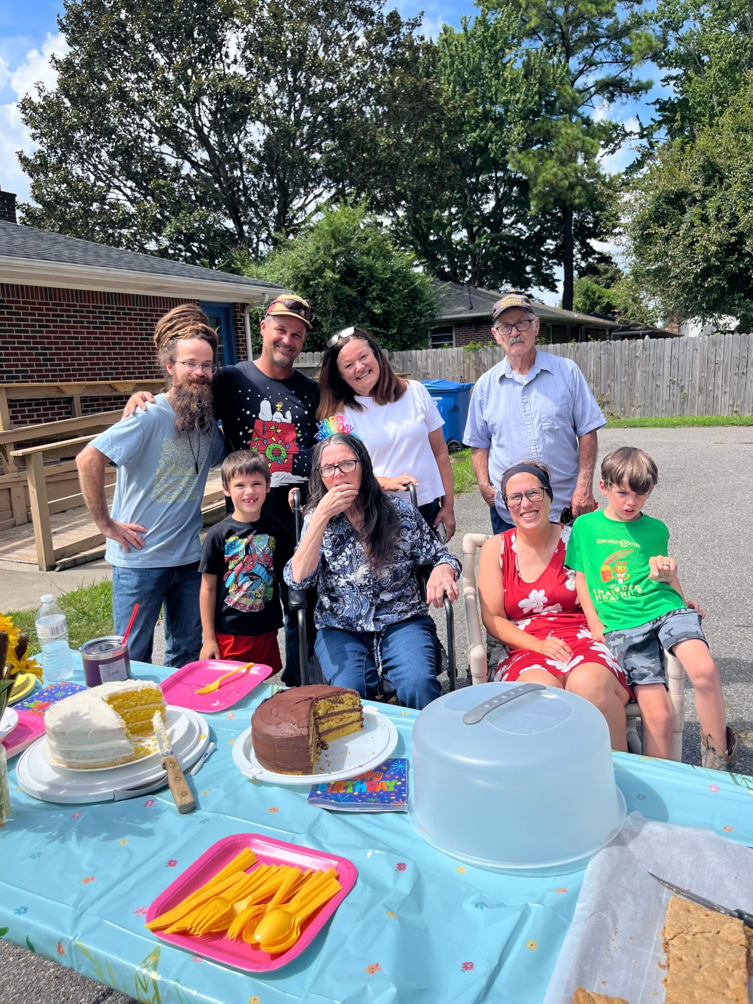 Group of people gathered outdoors around a table with food and drinks, celebrating a birthday with a cake and snacks.