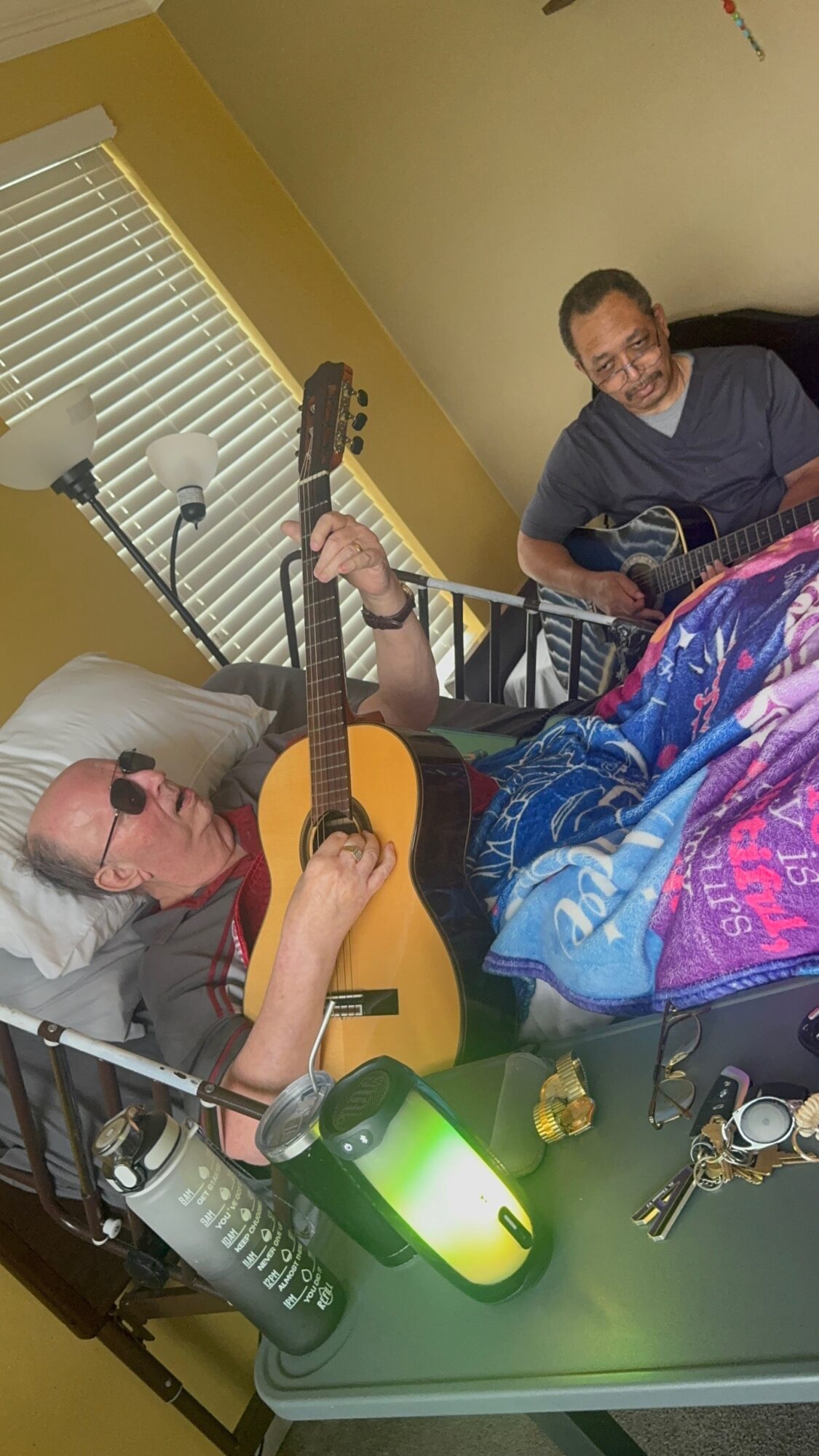 Two men in a room, one lying in bed with sunglasses and guitar, the other sitting nearby with a guitar, colorful blanket, and a green lamp.