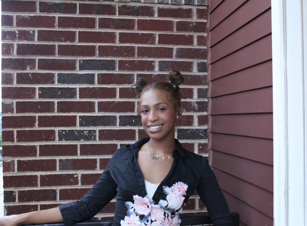 Young girl with two buns hairstyle smiling outdoors, standing in front of brick and siding wall.