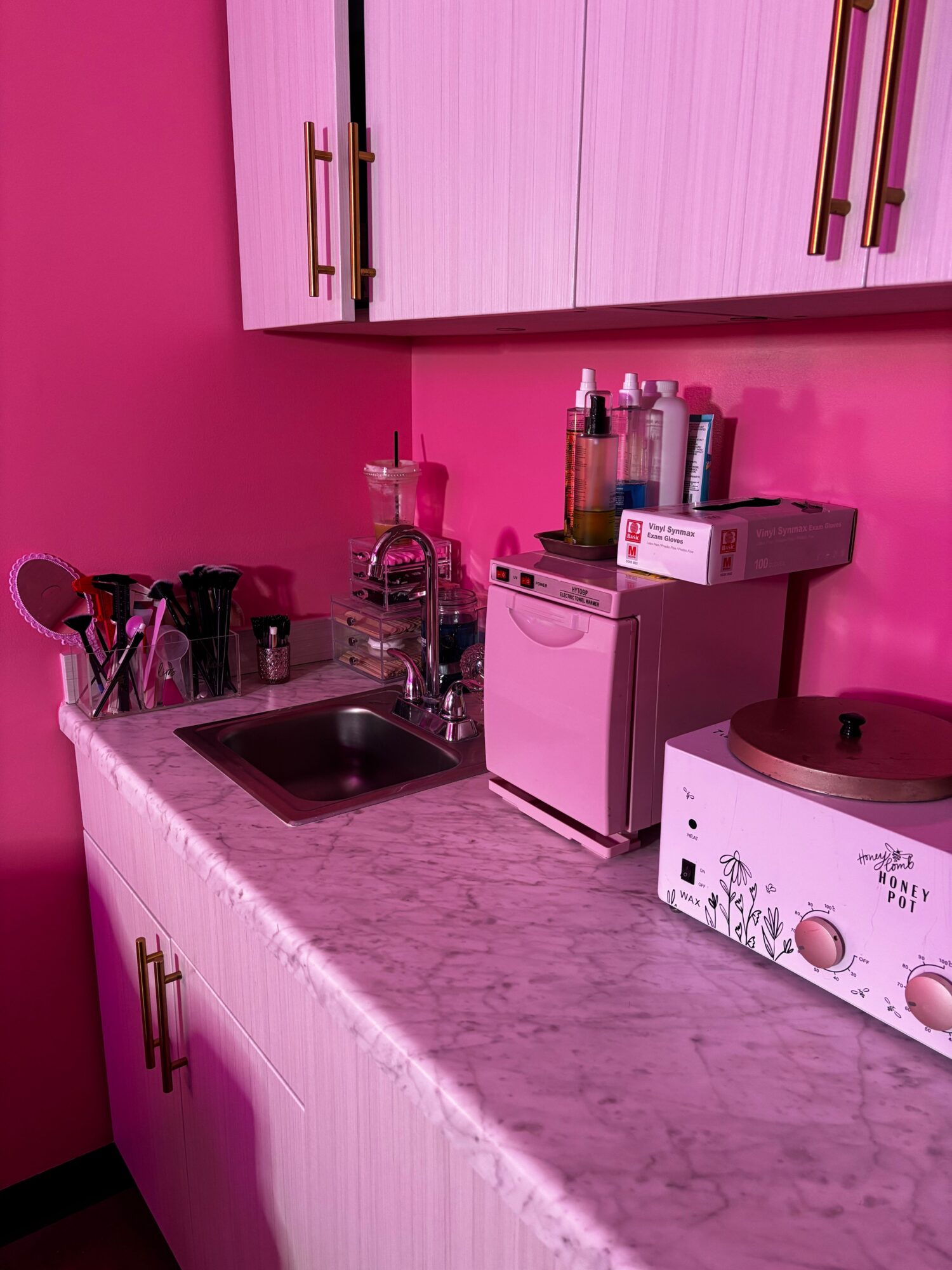 Pink kitchen with white cabinets, countertop, and various bottles and utensils, viewed from the side.