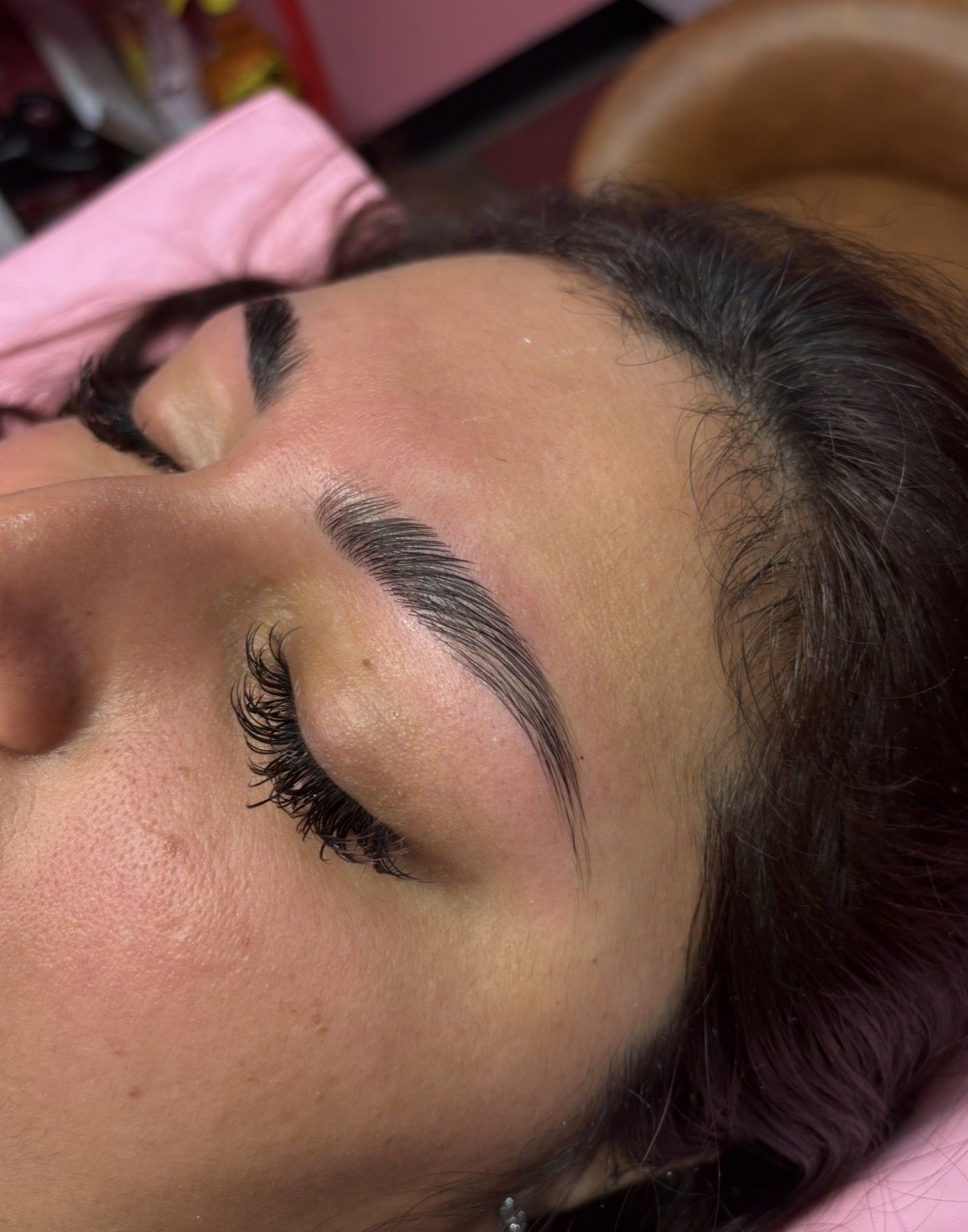 Close-up of a woman's face with eye closed, showing well-groomed eyebrows and long eyelashes, lying on a pink pillow.
