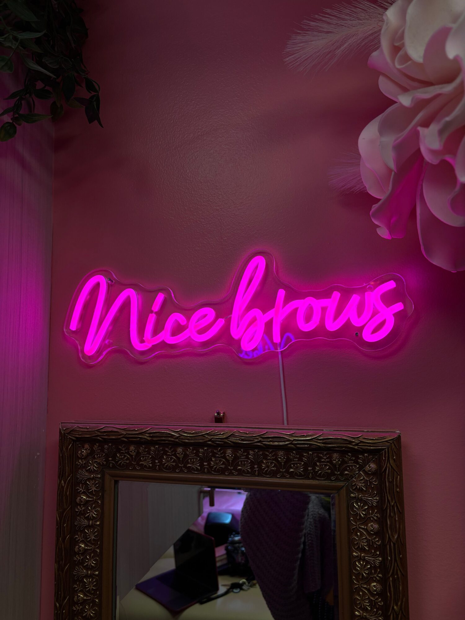 Pink neon sign reads 'Mischief' on a pink wall, with a mirror and decorative flowers nearby.