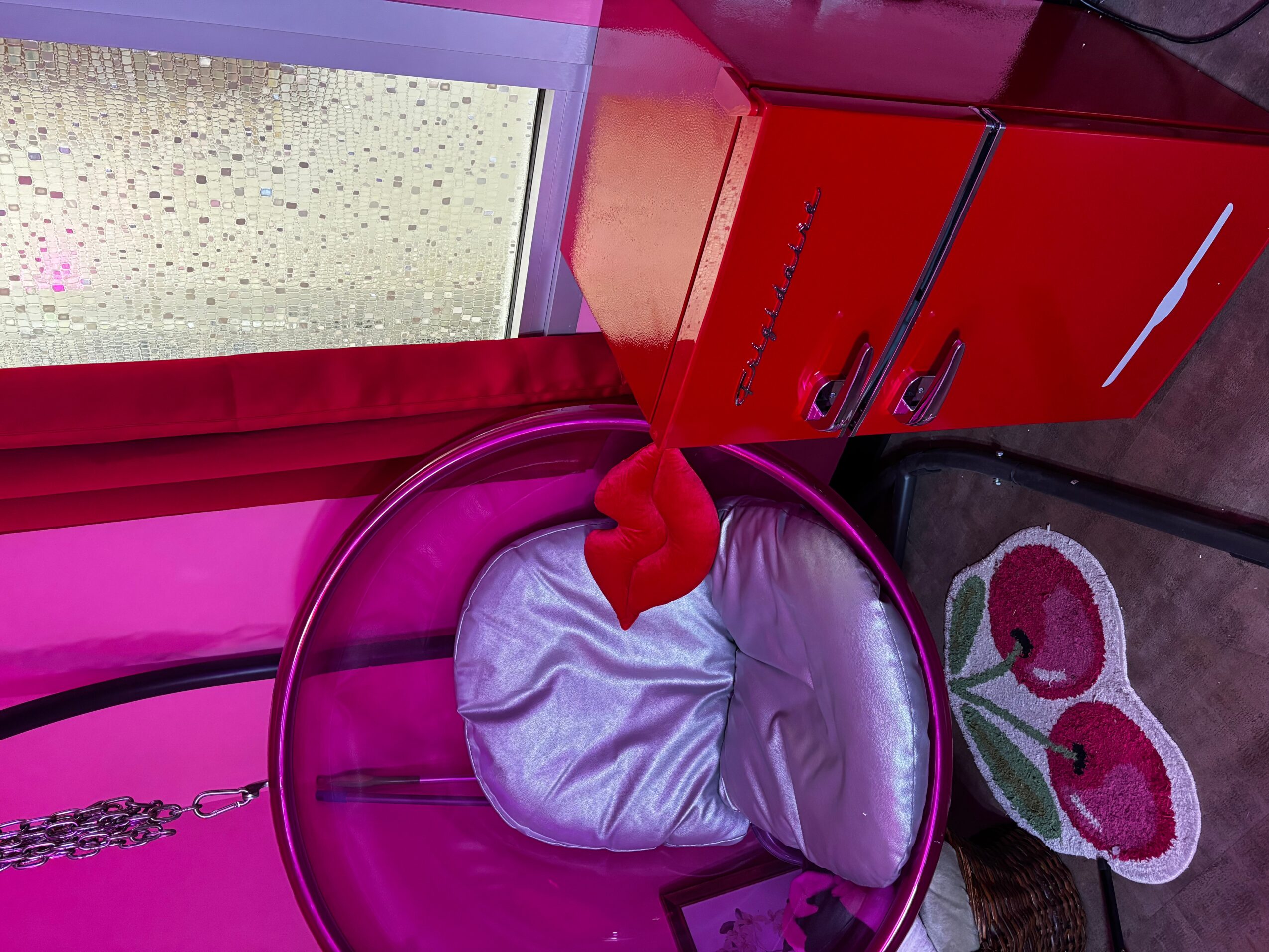 Pink laundry basket with a white cloth inside, red handle, pink walls, red cabinet, and a window with raindrops, colorful rug with cherries.