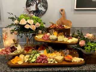 Assorted cheeses, fruits, and flowers on a kitchen counter with a cutting board and decorative items in the background.