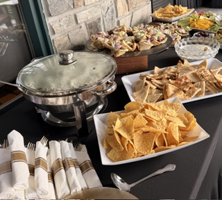 Buffet table with chips, sandwiches, and other snacks, set against a brick wall, with napkins and utensils in foreground.