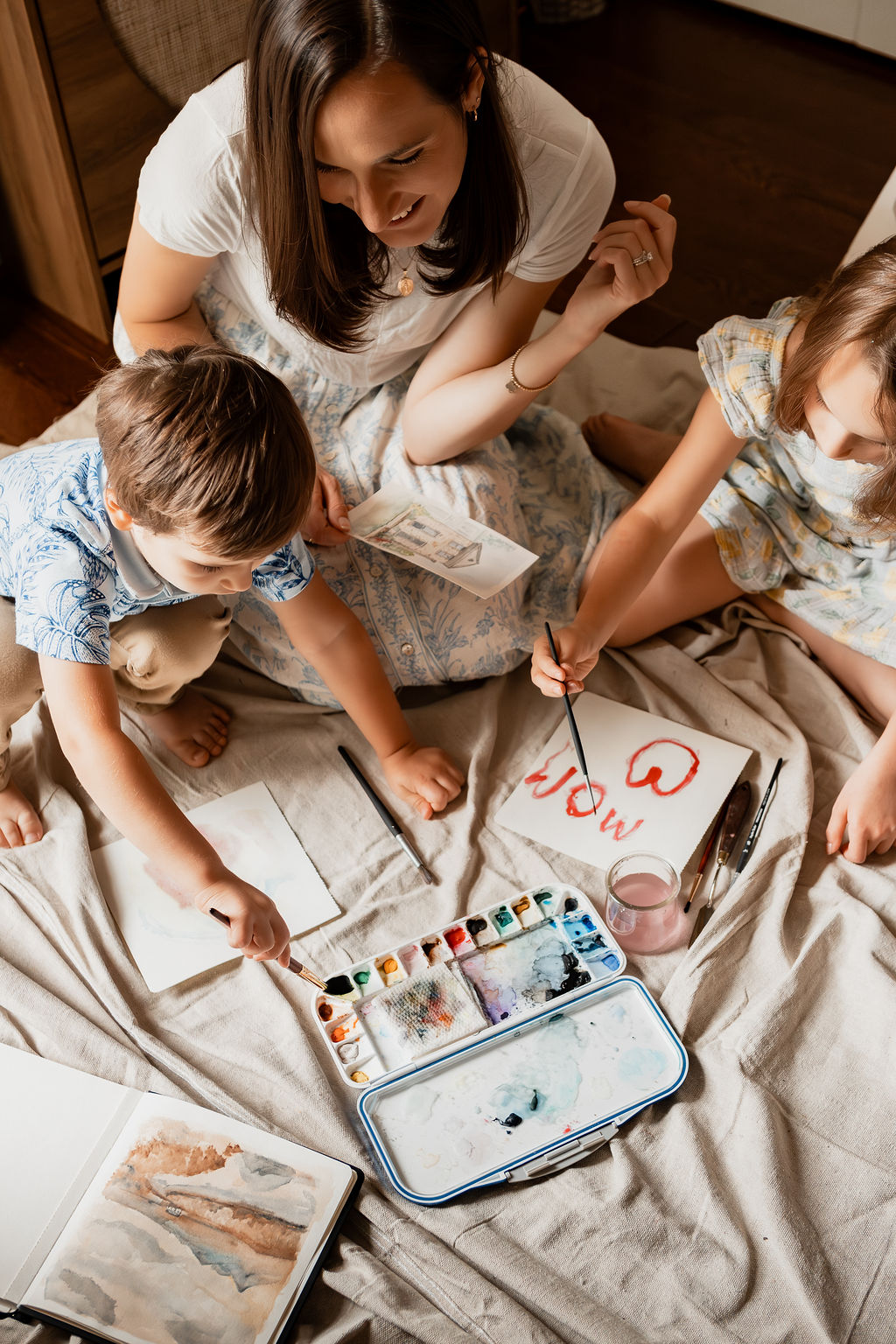 Woman and two children painting on paper with watercolors on a bed, viewed from above.
