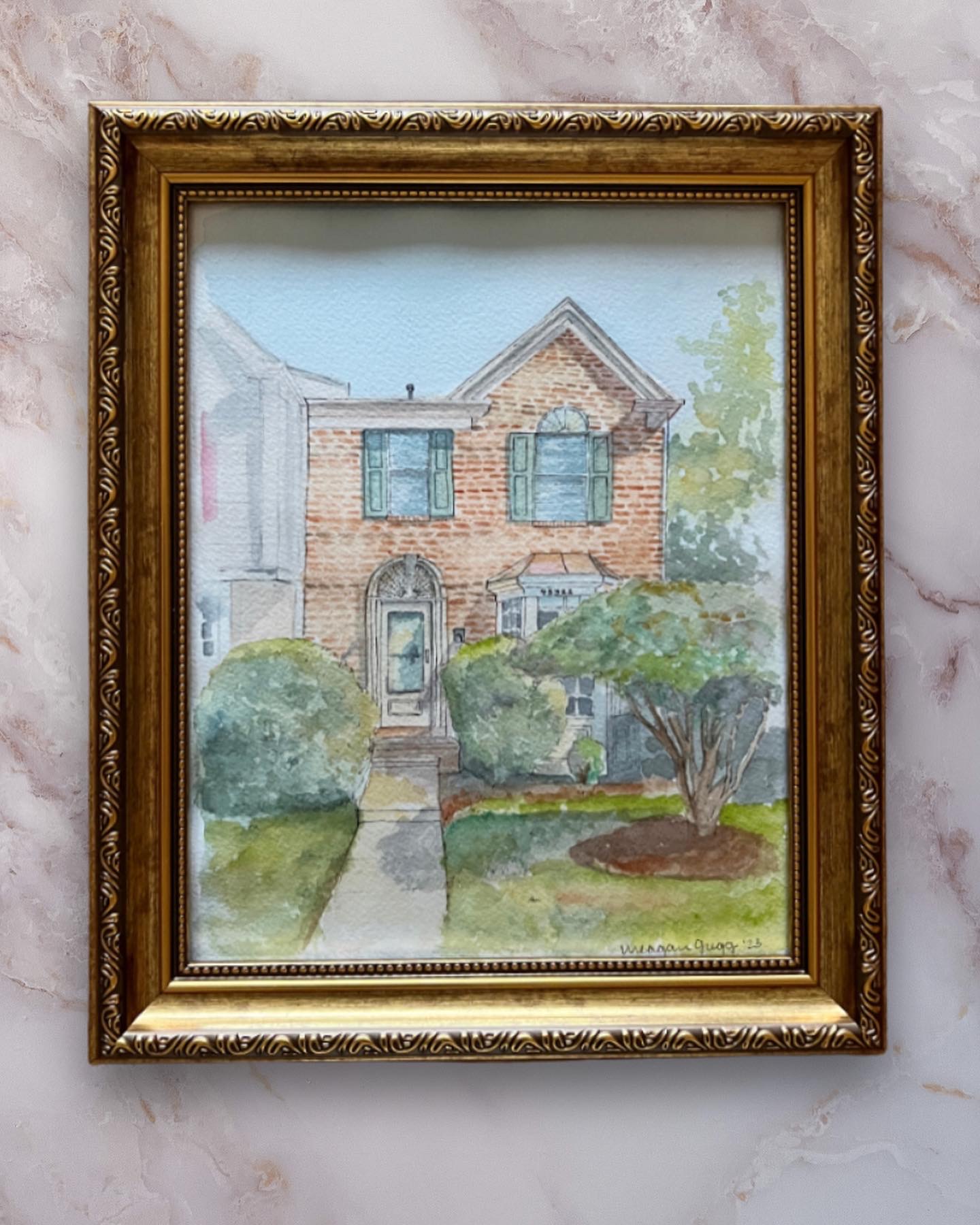 Watercolor painting of a brick house with trees and bushes in front, framed in an ornate gold frame.