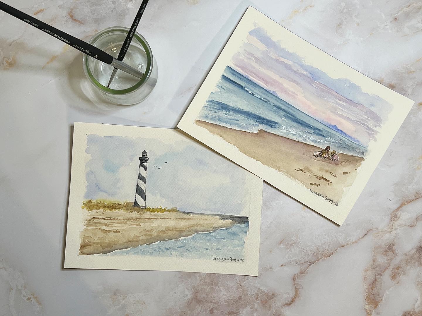 Two watercolor paintings of seaside scenes, one with a lighthouse and the other with a beach and ocean, on a marble surface.