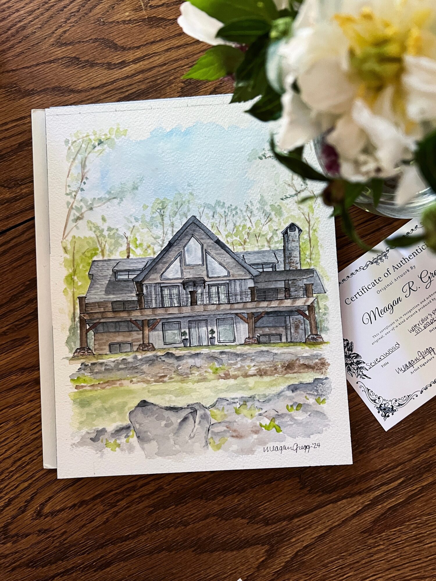 Watercolor painting of a house with a gabled roof, chimney, and porch, set against trees and sky, on a wooden surface.
