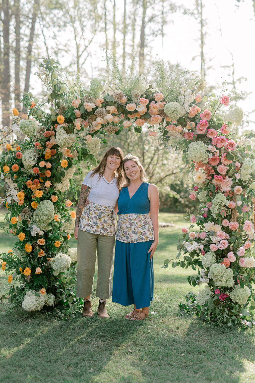 Two women stand under a floral arch outdoors, smiling, surrounded by trees and greenery.