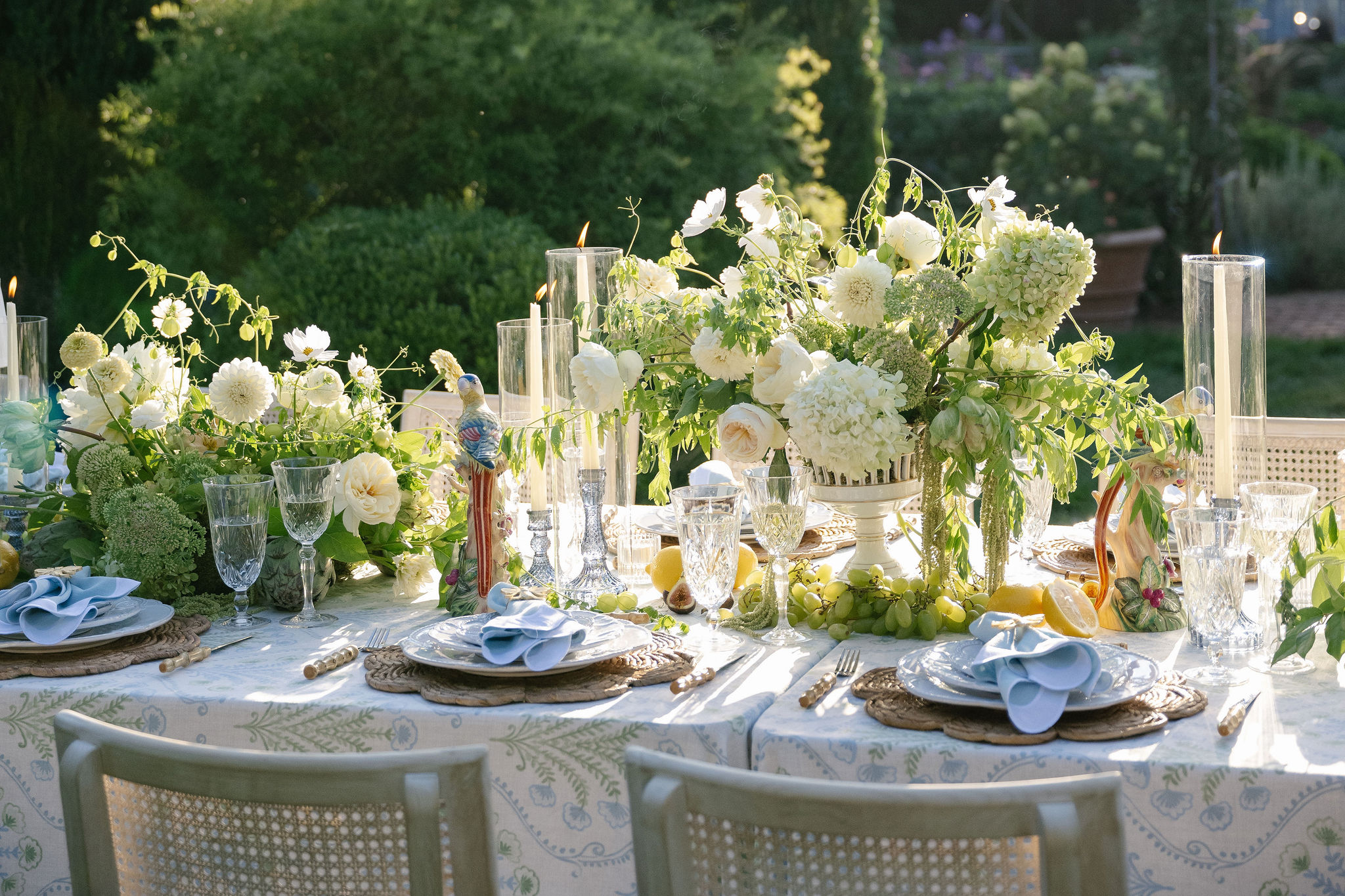 Elegant table setting with white flowers, glassware, and tableware outdoors, sunlight illuminating the scene.