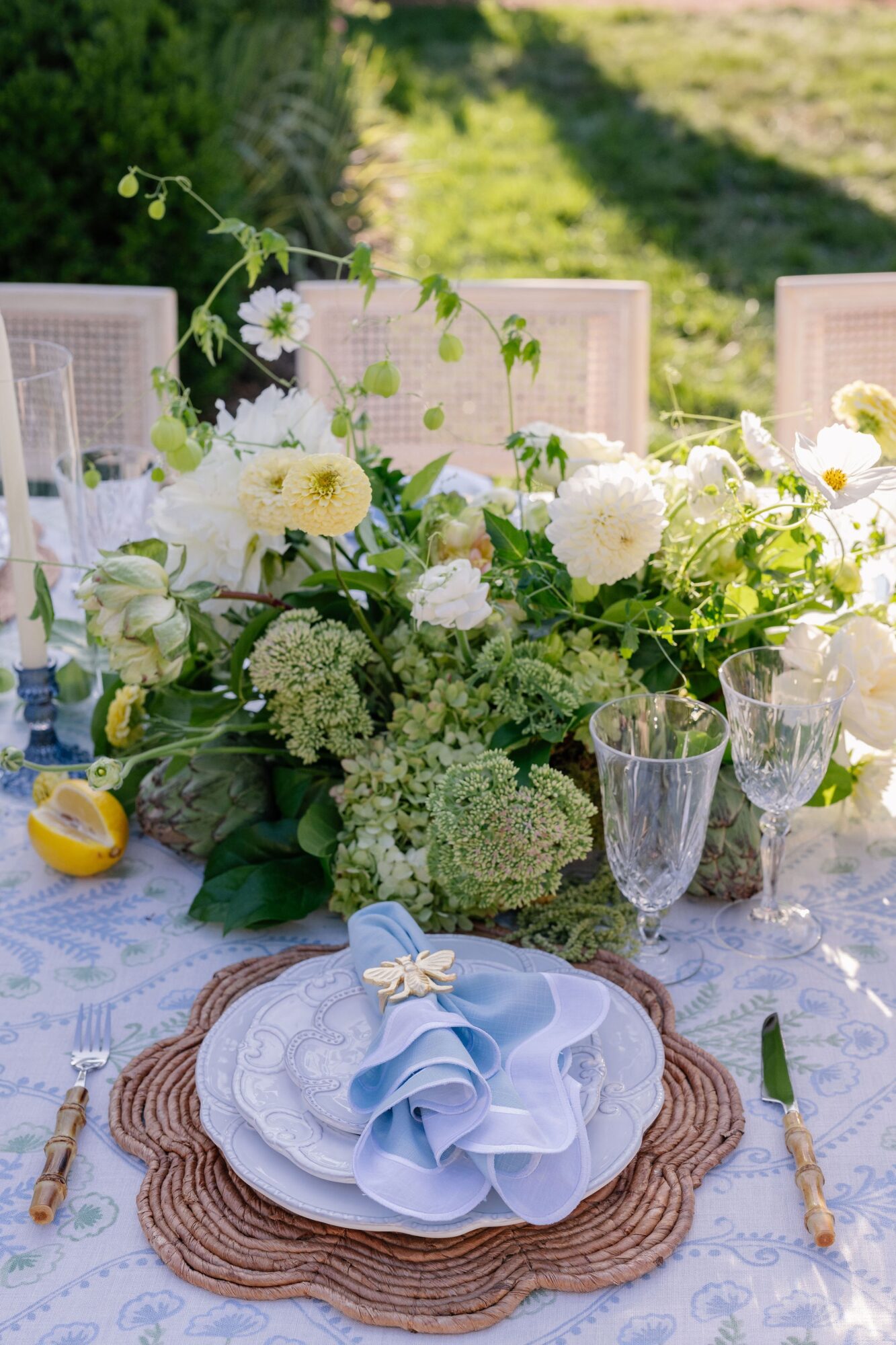 Table setting with white flowers, glassware, and a blue napkin on a woven placemat outdoors, sunny background.