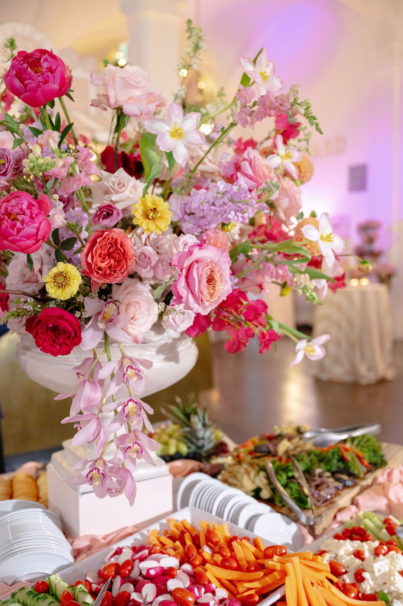 Large floral arrangement with pink, red, and yellow flowers in a white vase, with food and table setting in background.