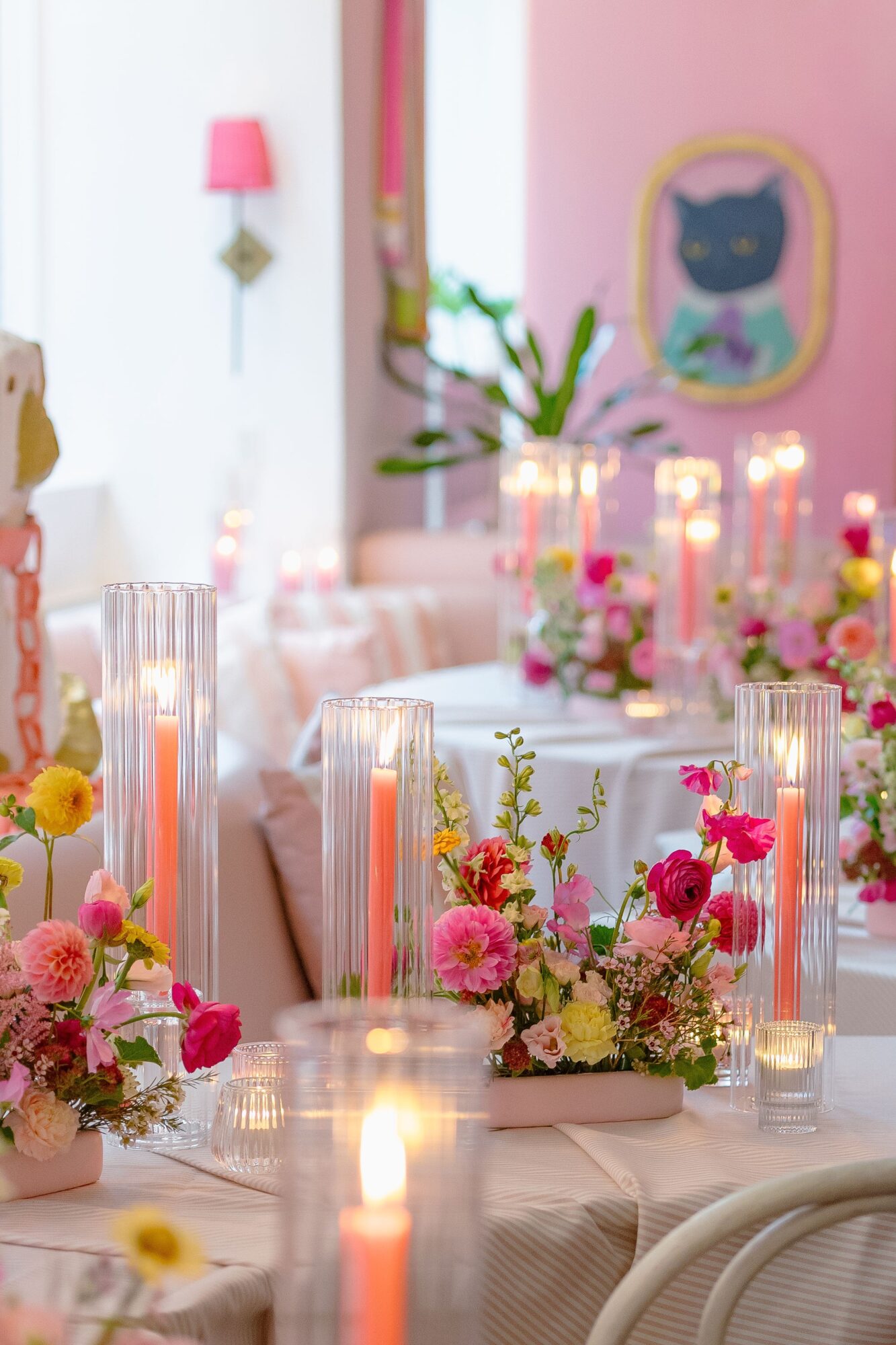 Decorated table with candles and floral arrangements in a pastel-colored room with pink walls and wall art.