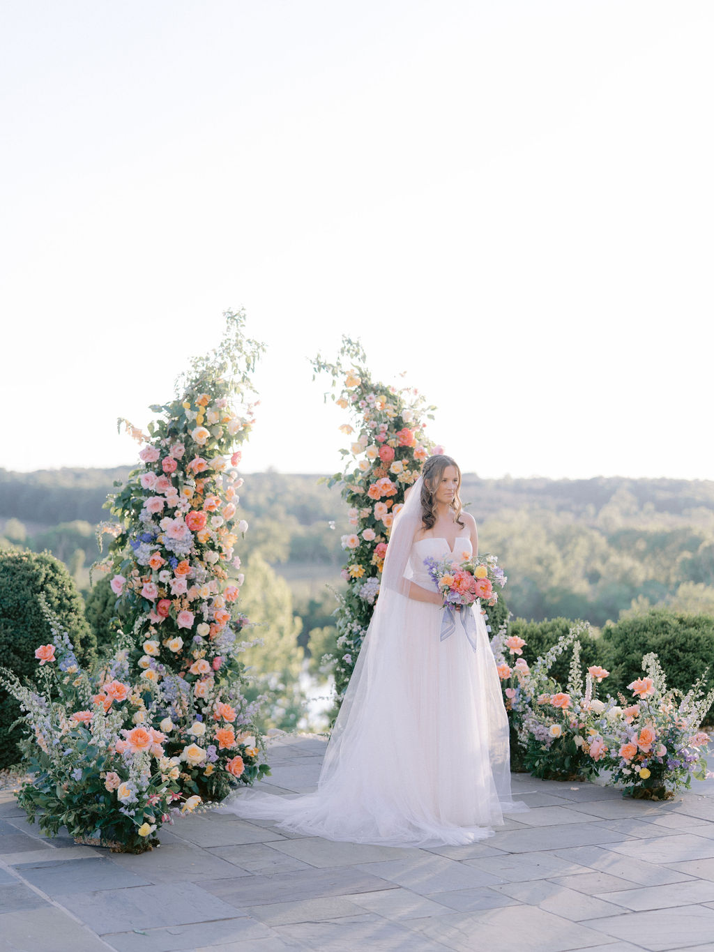 Bride in white dress holding a bouquet, standing outdoors near floral arch with greenery and sky in background.