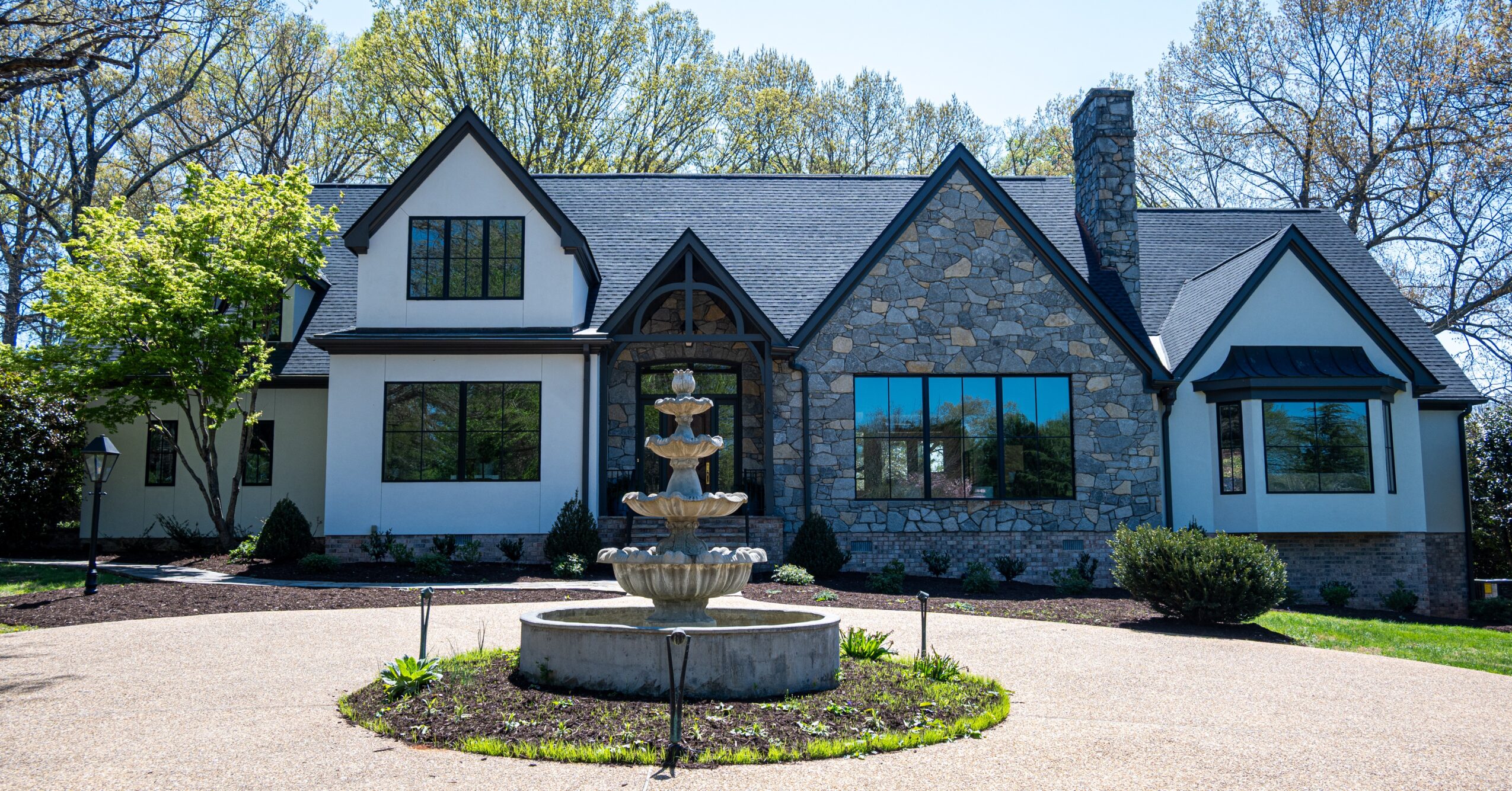 Large house with stone and white siding, front yard with fountain, trees, and clear sky.