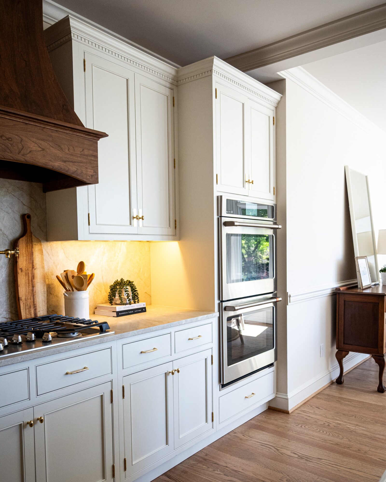 White kitchen cabinets with built-in oven, countertop with utensils, and a wooden floor, in a well-lit room.