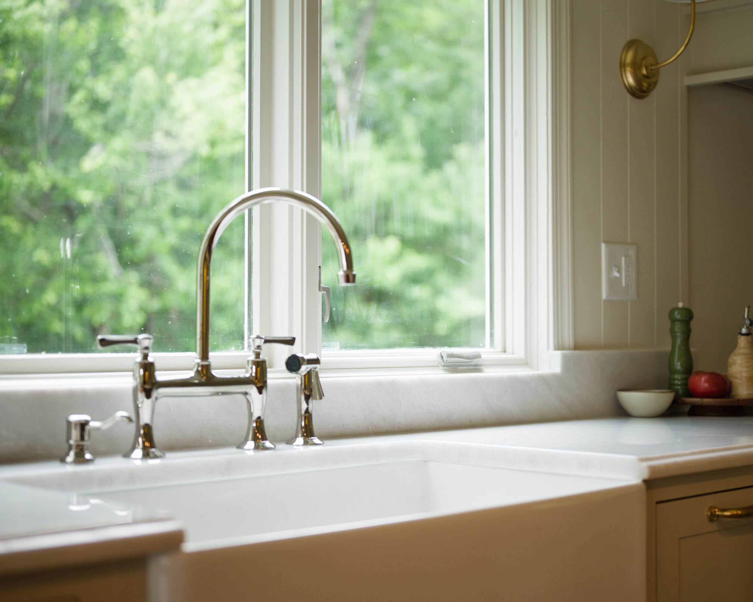 Kitchen sink with a window showing green trees outside, brass faucet, and countertop with small bowls and decorations.
