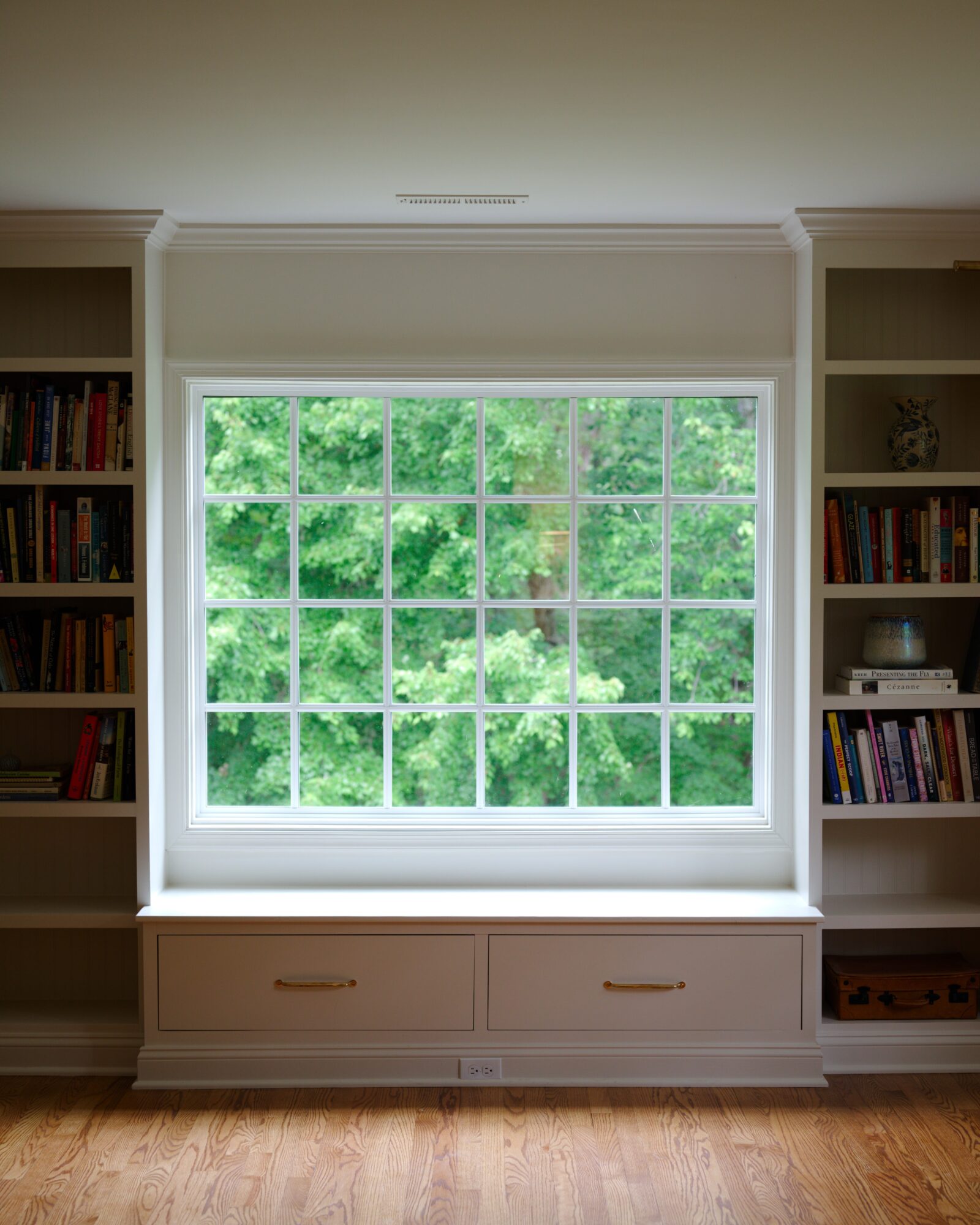 Large window with multiple panes showing green trees outside, flanked by built-in bookshelves on both sides, and a wooden floor.