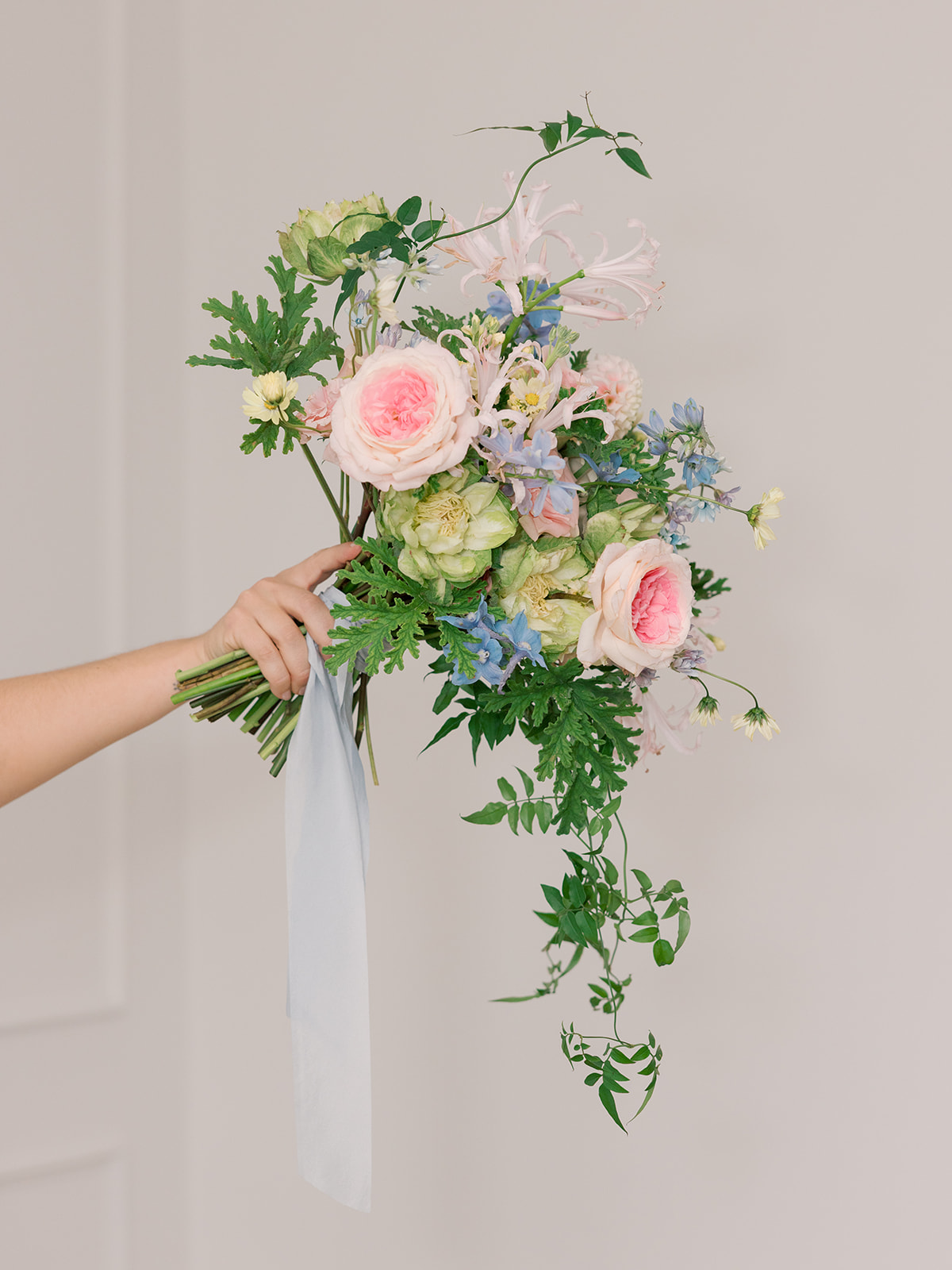 Person holds a cascading bouquet of pink, green, and blue flowers with greenery and a white ribbon.