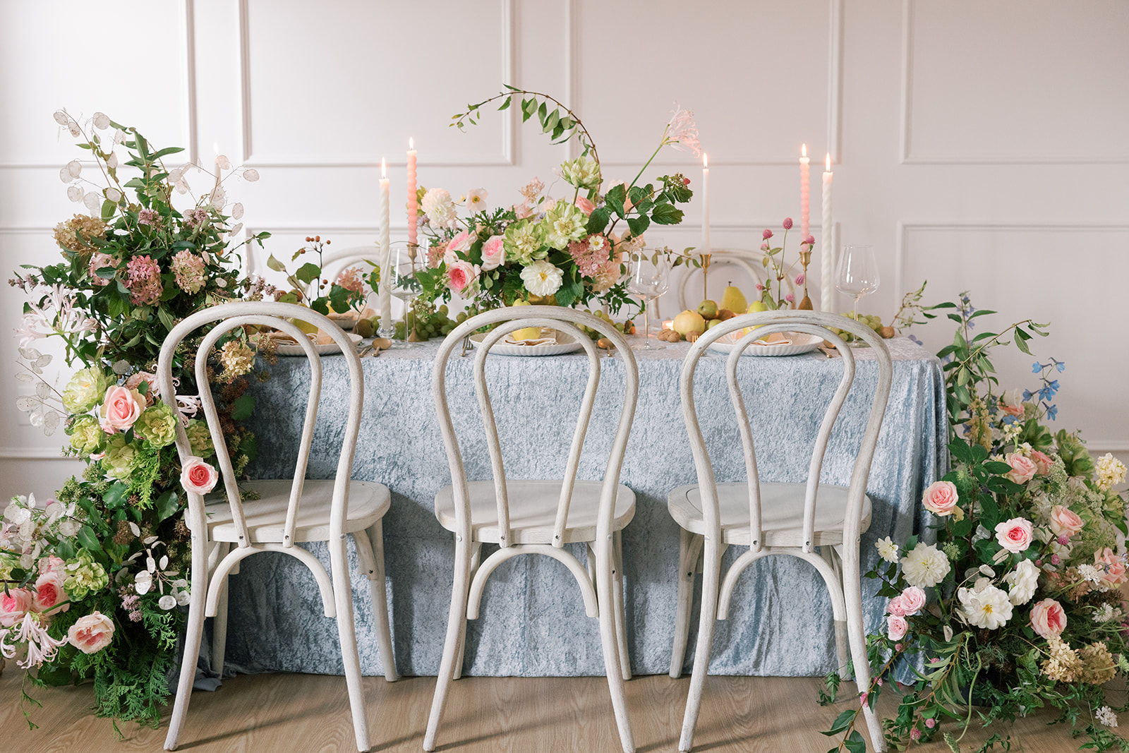 Table with floral arrangements, candles, and chairs in a decorated room, with a light blue tablecloth and pink and white flowers.