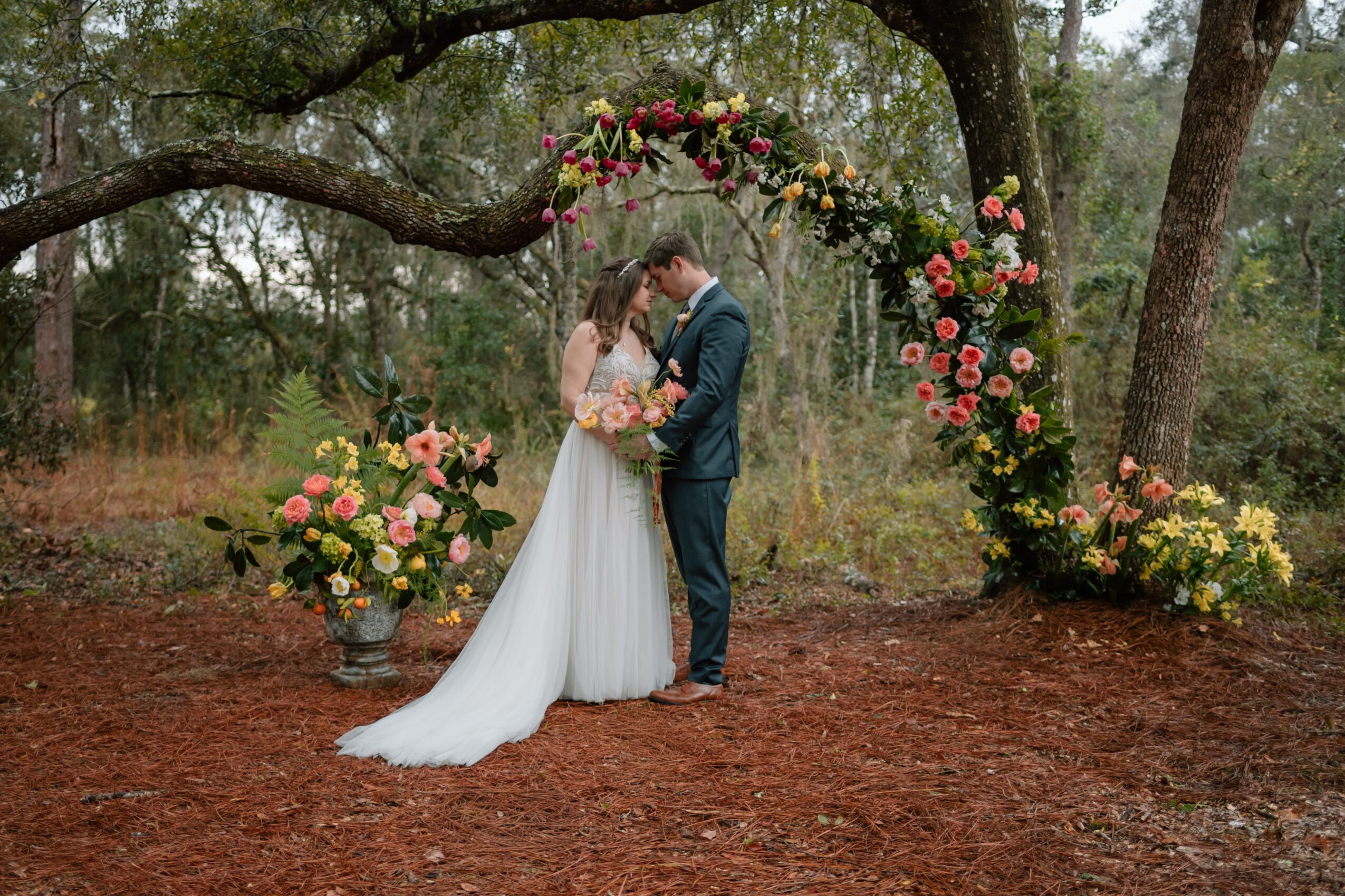 A bride and groom stand under a floral arch in a wooded outdoor setting, holding hands and looking at each other.