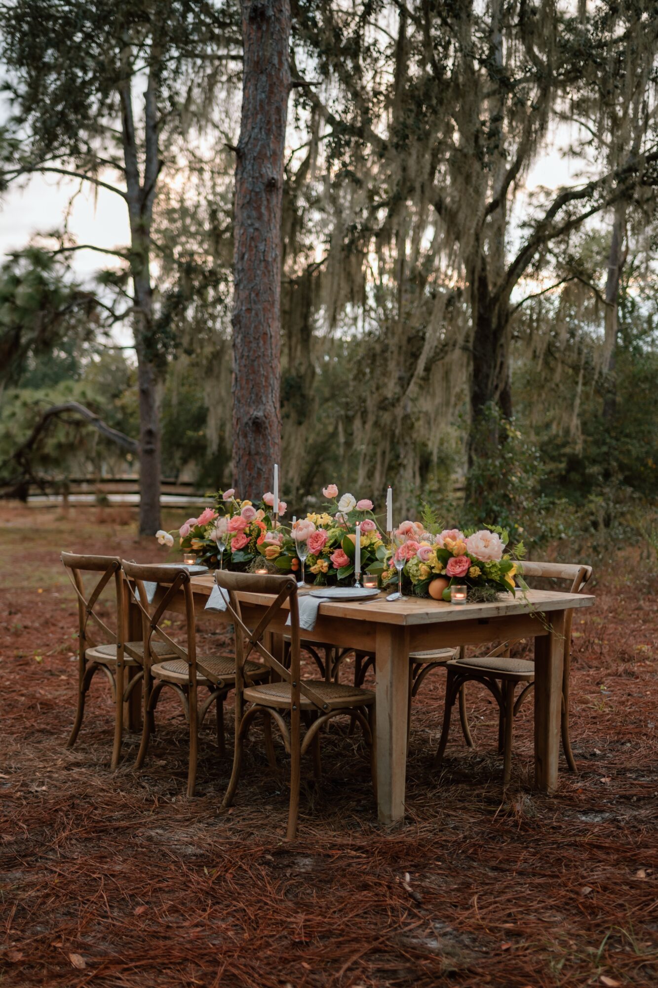 A wooden table with six chairs outdoors, decorated with pink and white flowers, in a wooded area.