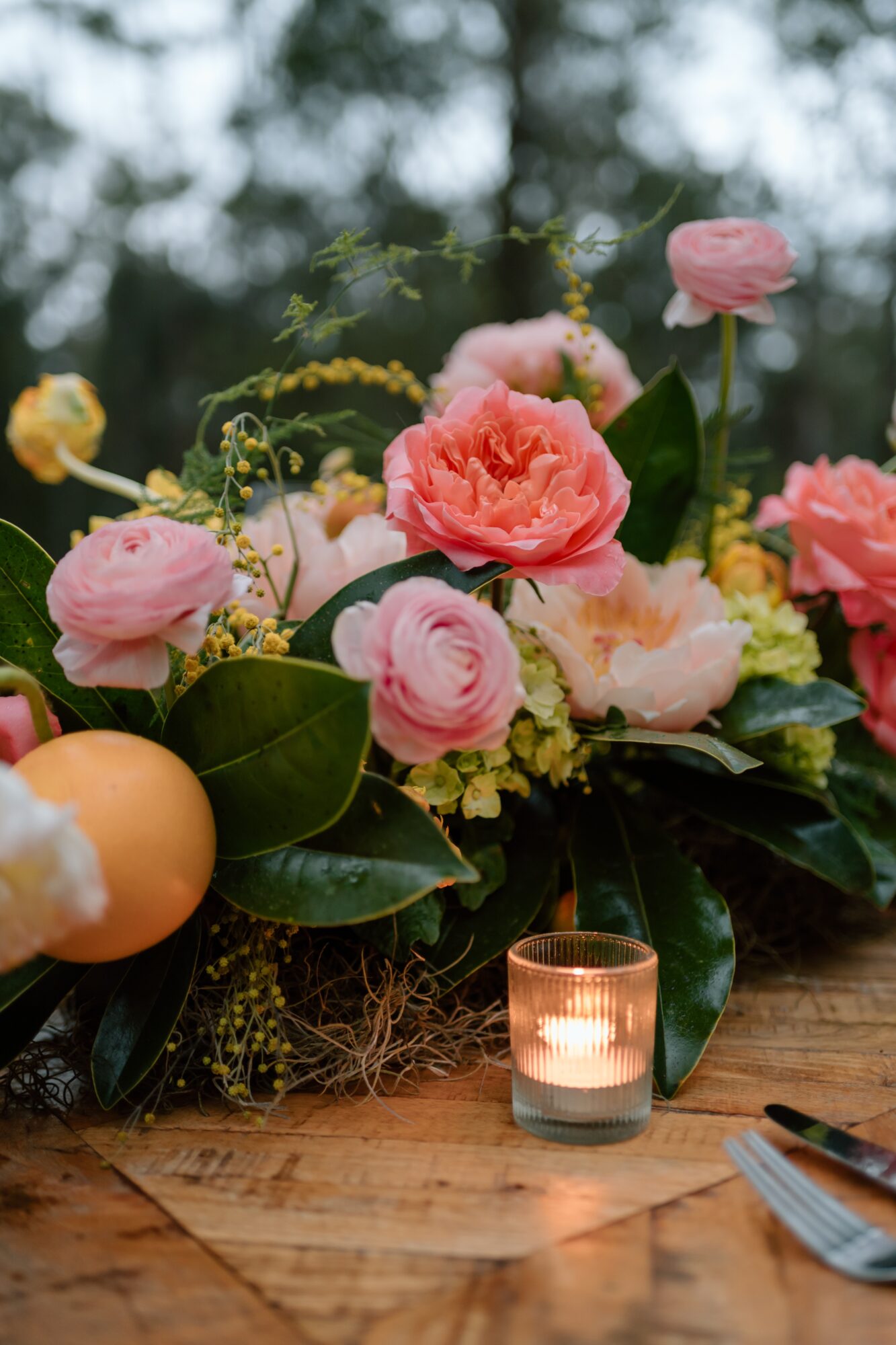 Pink and yellow flowers with green leaves and a lit candle on a wooden surface.
