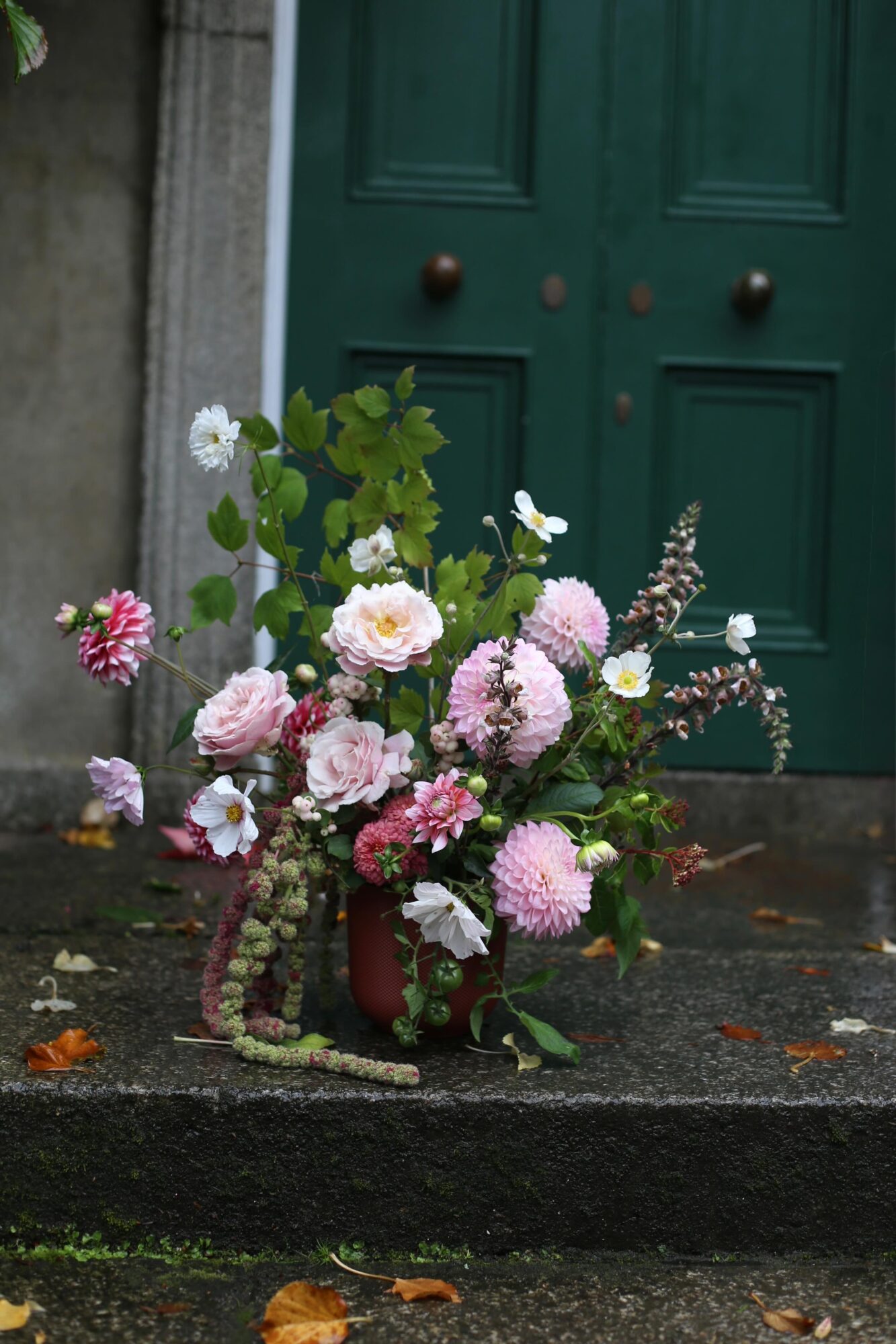 Flower arrangement with pink, white, and purple flowers in a red vase on a step in front of a green door.