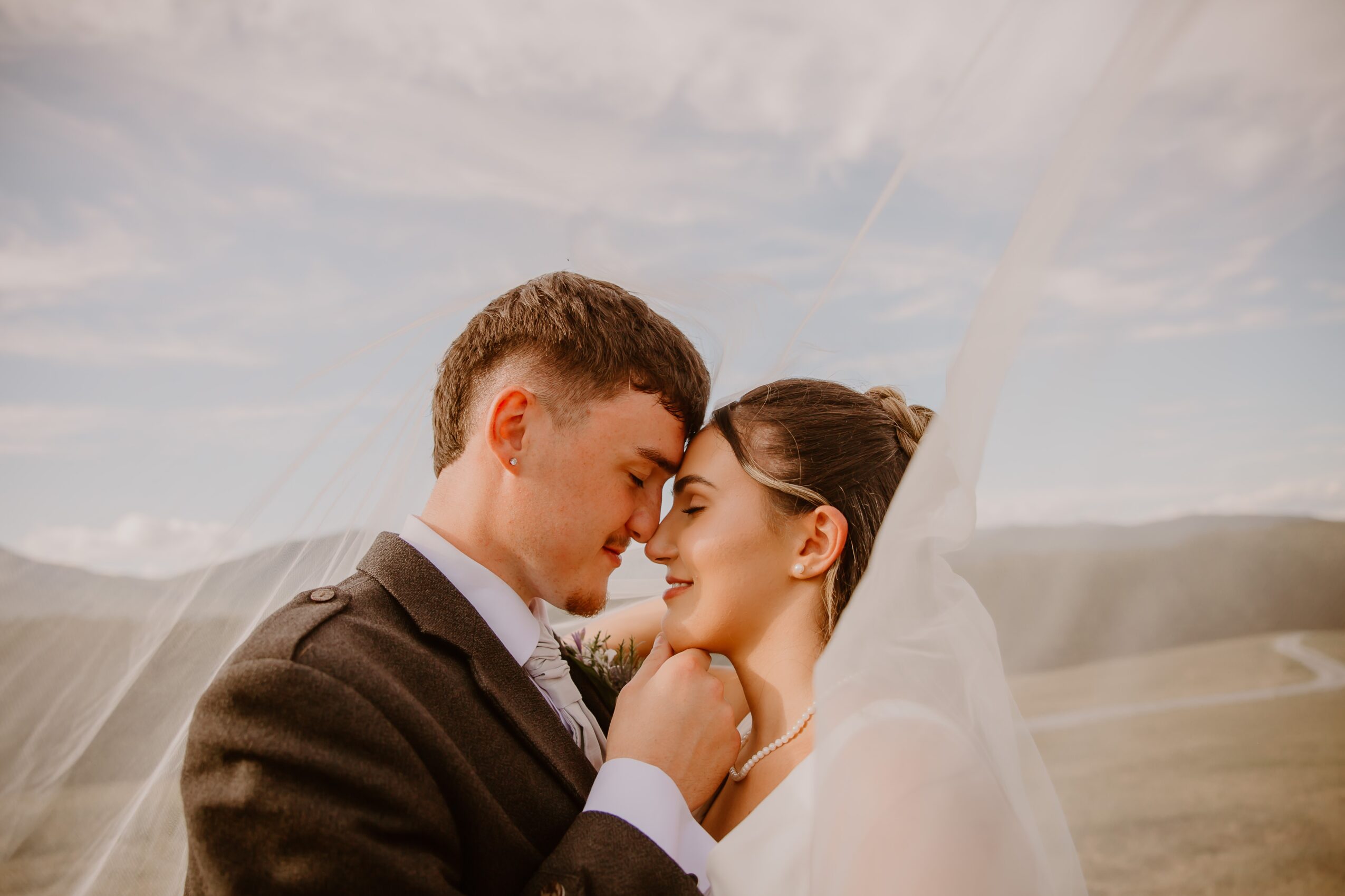 A couple in wedding attire face each other closely outdoors, with a scenic landscape and sky in the background.