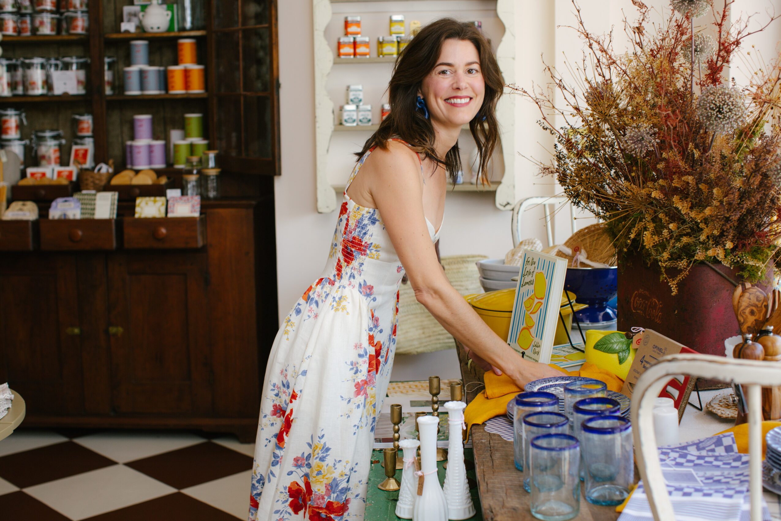 Woman in a floral dress smiling in a kitchen with shelves, jars, and a large flower arrangement on the counter.